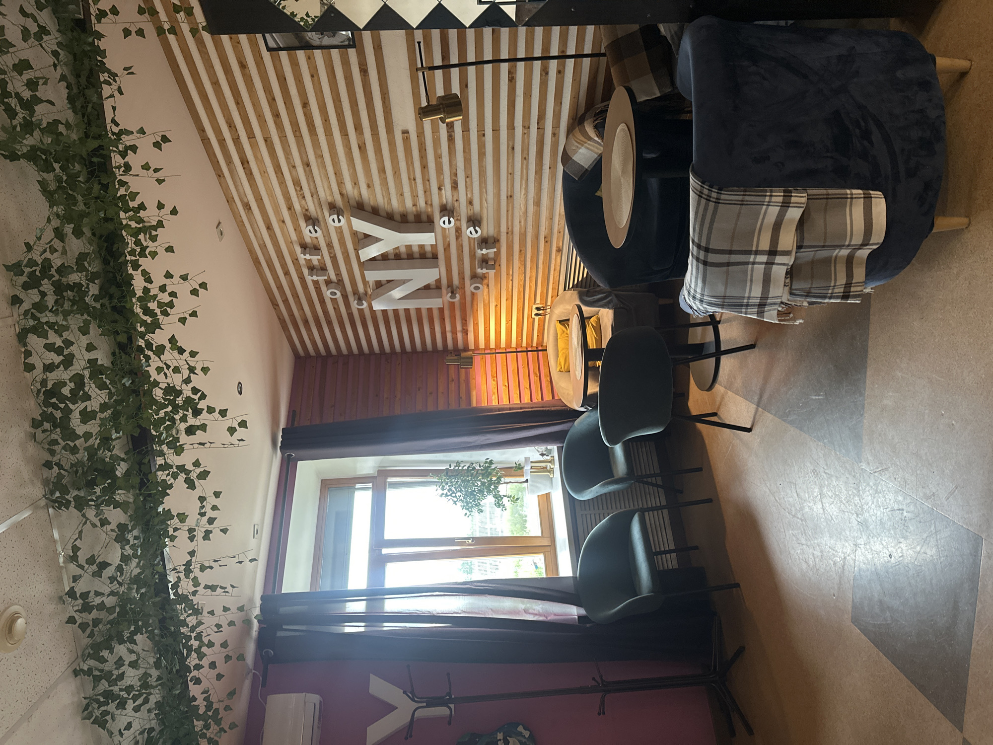 This is a photograph of the interior of a cafe. The cafe has a modern design with wooden slat walls and a ceiling decorated with artificial ivy. There are several seating areas with comfortable chairs and small tables. One wall features large white letters spelling out "NY" with "coffee" written around it. Natural light streams in from a window. The floor is a patterned linoleum. The image appears to have been taken in Votkinsk, Russia, given the context provided.