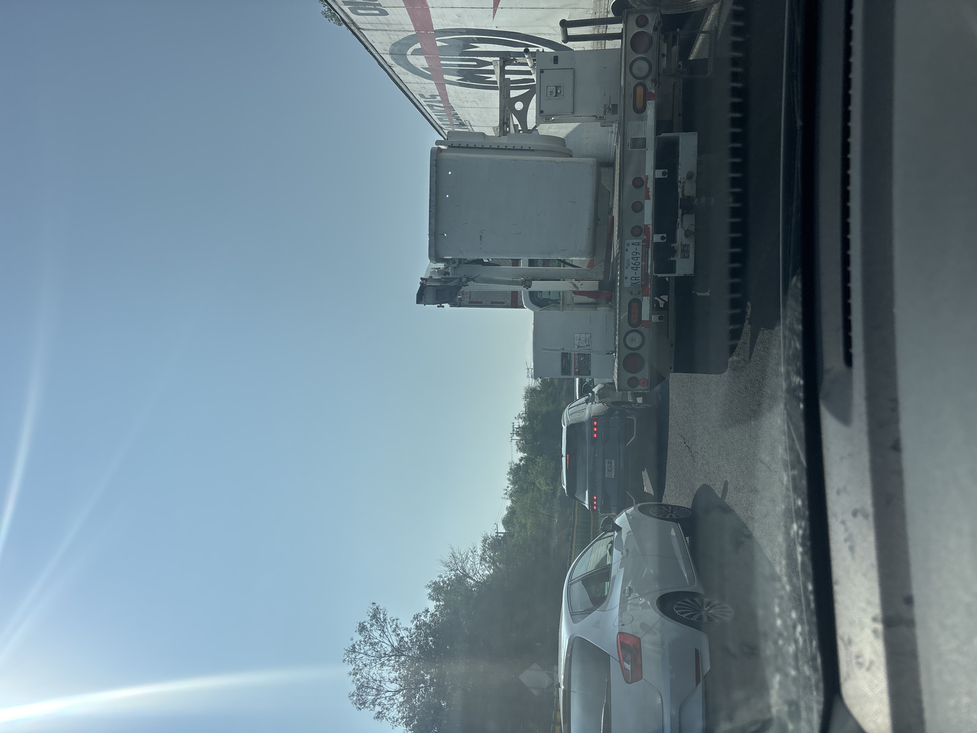 The image shows a view from inside a vehicle, likely a car, looking out at traffic. A large utility truck with a bucket lift is in the lane beside the camera vehicle. A gray car and a dark-colored van are visible in the distance behind the truck. The sky is light blue, and trees are visible along the roadside. The location is Cuautitlán Izcalli, Mexico. The image depicts a traffic situation on a sunny day.