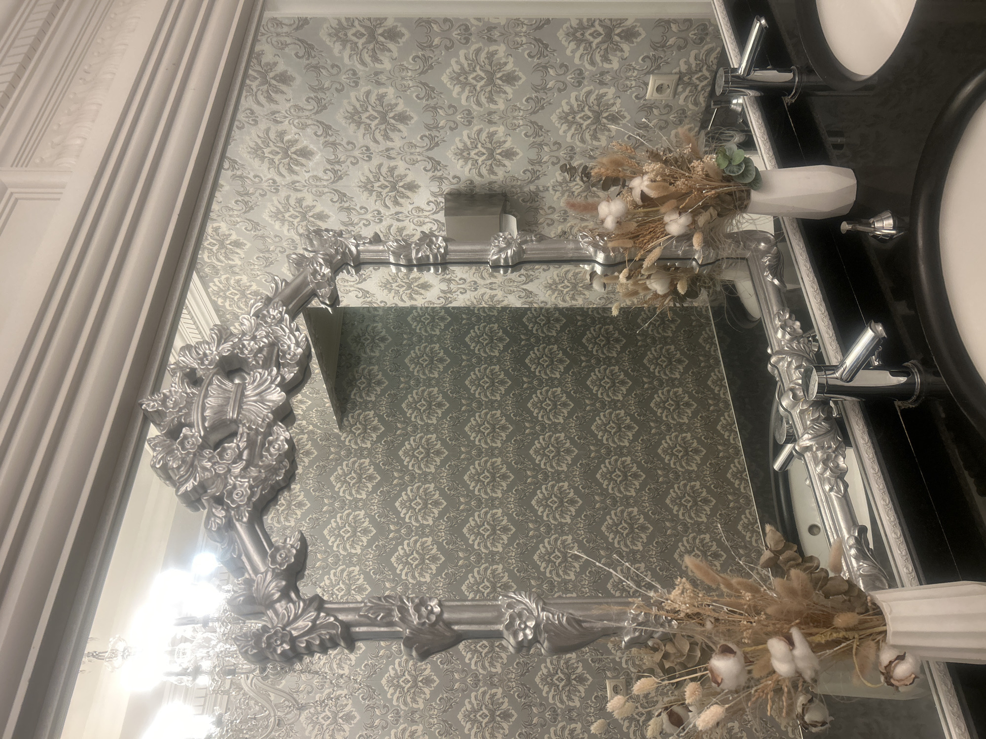 The image shows a close-up view of a bathroom vanity area in Almaty, Kazakhstan. The vanity features two sinks, each with a chrome faucet. A large, ornate silver mirror with intricate floral and scrollwork details dominates the upper portion of the frame. The mirror is framed by decorative molding on the wall above. The wall is covered in a damask-patterned wallpaper in shades of gray and silver.
In the foreground, on the black granite countertop, are two white vases containing dried floral arrangements. These arrangements include elements like cotton bolls, dried grasses, and other natural fibers, some with metallic accents. To the left of the arrangement, a chrome soap dispenser is visible. A reflection of a chandelier and the patterned wallpaper is seen in the mirror. Electrical outlets are visible on the wall behind the vanity. The overall impression is one of a luxurious and elegantly decorated bathroom.