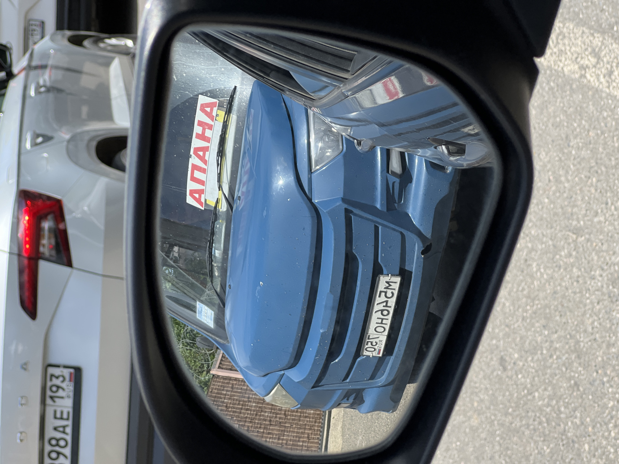 The image captures a reflection in a car's side mirror on a bright, sunny day, likely in Krasnodar, Russia.
The primary subject reflected in the mirror is the front of a blue car or van, appearing dusty. On its windshield, a red-lettered sticker is visible, which, when un-reversed from the mirror's reflection, reads "АНАПА" (Anapa, a city in Russia). The blue vehicle also displays a Russian license plate on its front. Reflected in the mirror, the plate shows "020 OHO 93 RUS", which would correctly be "OHO 020" for the main registration and "93 RUS" for the region code, indicating Krasnodar Krai.
Behind the blue vehicle, and outside the mirror's direct reflection but visible in the background, is the rear of a white car. This car also has a Russian license plate clearly visible: "B98AE 193 RUS". The "193" region code also corresponds to Krasnodar Krai. The white car's red taillights are partially visible.
The scene depicts parked or stationary vehicles on an asphalt surface, with what appears to be a building or fence structure reflected subtly in the background of the mirror. No specific activity or people are shown, only the static arrangement of cars. The strong sunlight suggests it is daytime.
