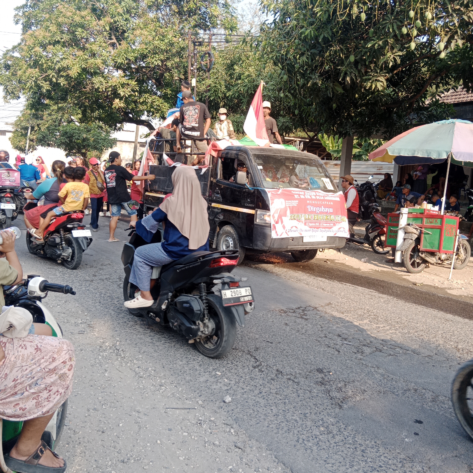 In an outdoor street scene, possibly in Tegalrejo, Indonesia, a procession is underway, likely celebrating a national holiday given the Indonesian flag prominently displayed. A black truck, decorated with a large red and white banner proclaiming "Mengucapkan Dirgahayu REPUBLIK INDONESIA KE-," is the central focus. The banner also includes text about unity for a prosperous future. Several people are visible on and around the truck, some holding flags and looking towards the viewer or the side.
The street is paved and appears to be in moderate condition with some cracks and stones. On the right side of the frame, a food cart with "OJEK SIOMAY" written on it is visible, suggesting a public gathering with food vendors. Several motorbikes are present, with some individuals riding them and others parked. In the foreground, a person on a motorbike is partially visible, their patterned skirt and sandals indicating everyday attire. The background features trees with lush green foliage, some of which bear green fruit, and buildings with tiled roofs. The lighting suggests it is daytime, possibly late afternoon, with shadows cast on the road. The overall atmosphere appears festive and communal.