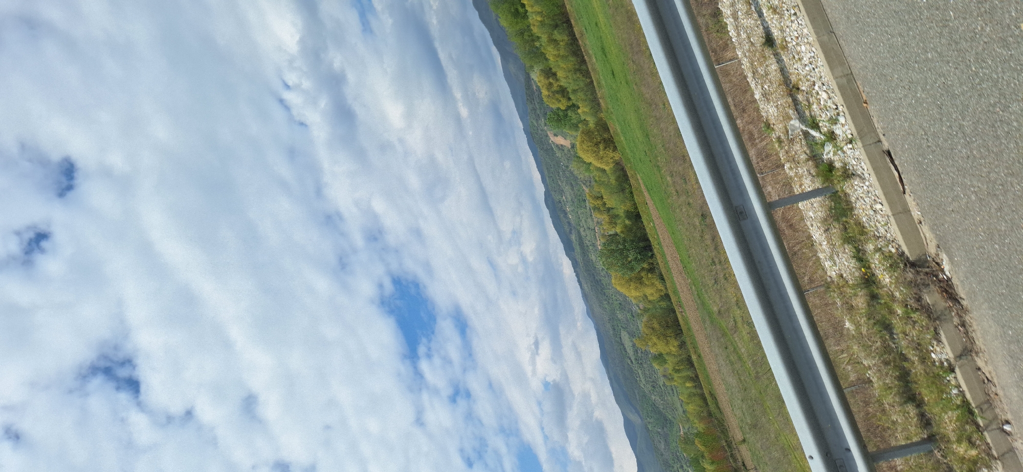 The image captures a scene along a highway near Valkovo, Bulgaria. In the foreground, a gray asphalt road is visible with a concrete curb and gravel shoulder. A silver metal guardrail runs parallel to the road, separating it from a green field. Beyond the field, a wooded hillside rises, its trees showing variations in color. The sky is filled with white clouds, interspersed with patches of blue. There are no visible people or actions in the scene. The focus is on the landscape and infrastructure.