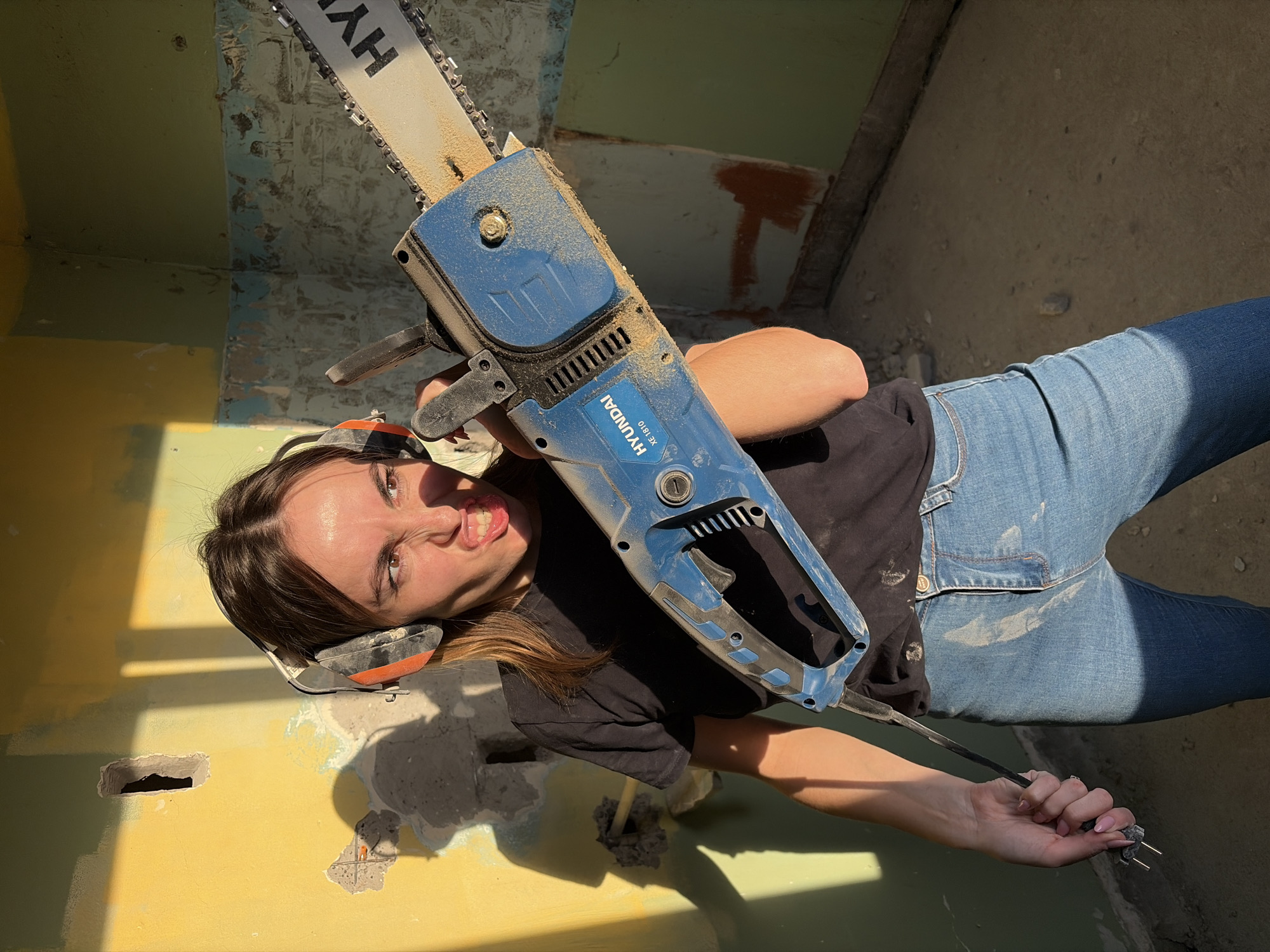 A woman wearing jeans, a black T-shirt, and earmuffs is holding a blue Hyundai XE 1810 electric chainsaw. The woman is squinting at the camera with her mouth open, showing her teeth. She is standing in a room with yellow and blue walls that appear to be under construction or demolition. There is a hole in the wall, and pieces of the wall are missing. The location appears to be Izhevsk, Russia, although this cannot be confirmed from the visual information alone.