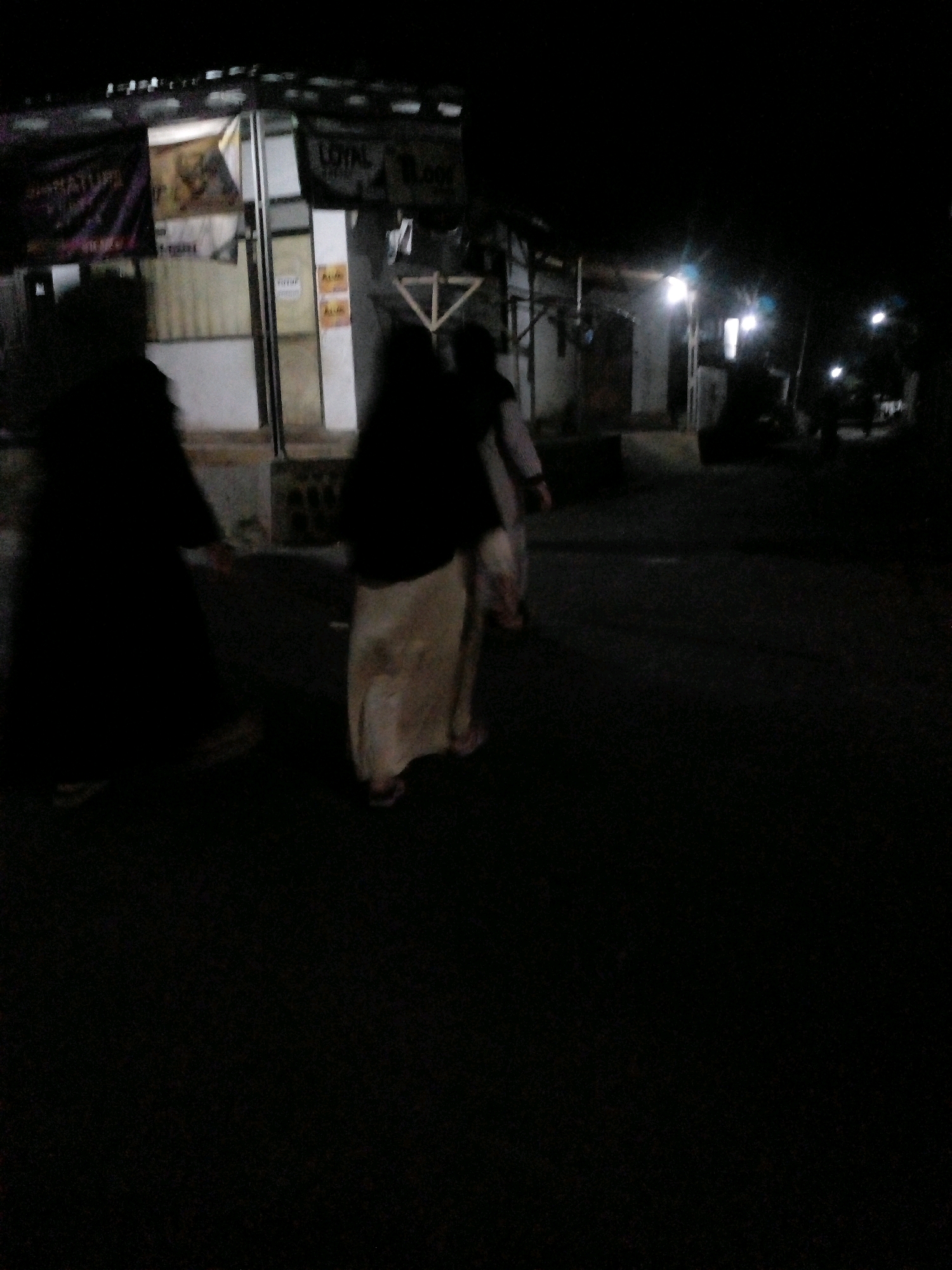 The image shows a street scene at night in Pungangan, Indonesia. Two individuals, appearing to be women, are walking away from the camera on the right side of the frame. The person in the foreground is wearing a dark abaya and a light-colored skirt, while the person behind them is wearing a dark abaya and a light-colored skirt. To the left, a person is silhouetted against a lit building. The building appears to be a shop or storefront with signage that is partially visible and illegible due to the low light conditions and image blur. The street is paved and dark, with some artificial lighting from street lamps visible in the distance, casting a glow on the scene. The overall atmosphere is dim and somewhat mysterious, typical of a street at night.
