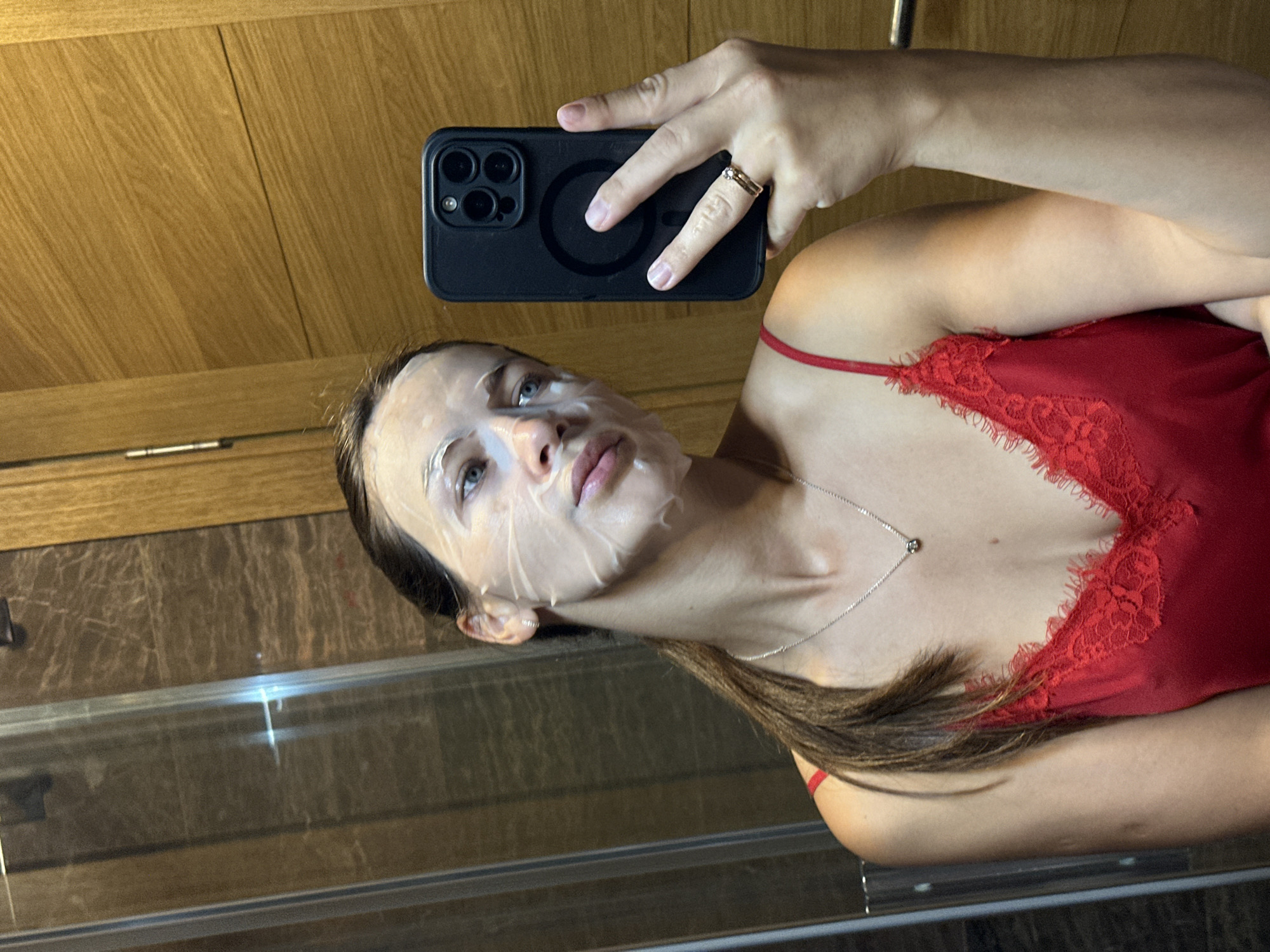 The media file is a selfie taken by a young woman with a face mask on. She is wearing a red camisole with lace trim and a silver necklace. Her hair is dark brown and pulled back. She is holding a black smartphone in her right hand, which is also wearing a ring. The background appears to be a bathroom with wooden paneling and marble tiles. The lighting suggests it is daytime. The woman's expression is neutral, and she is looking at the camera. No text is visible in the image. The location context provided is Donji Pobori, Montenegro.