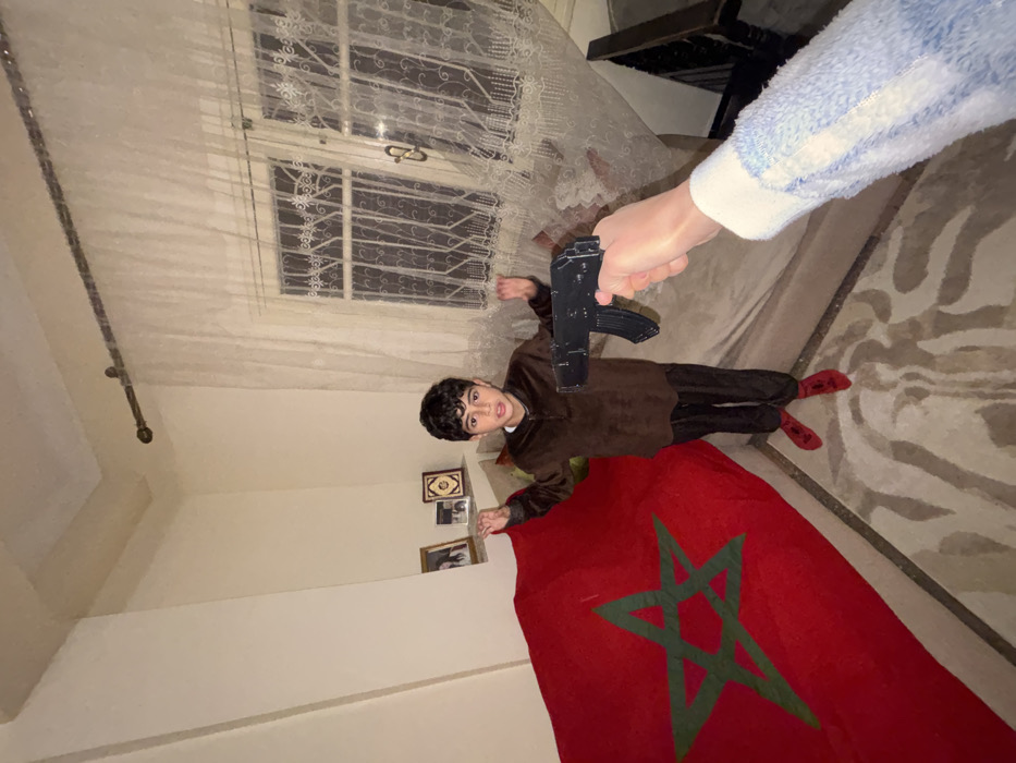 A young boy lies on a rug with a Moroccan flag beneath him. He is wearing a brown shirt and pants with red shoes. An arm extends towards him holding a black toy gun pointed at the boy. The scene takes place indoors, with a window covered by a white lace curtain visible. There are several framed pictures on the wall beside the boy.