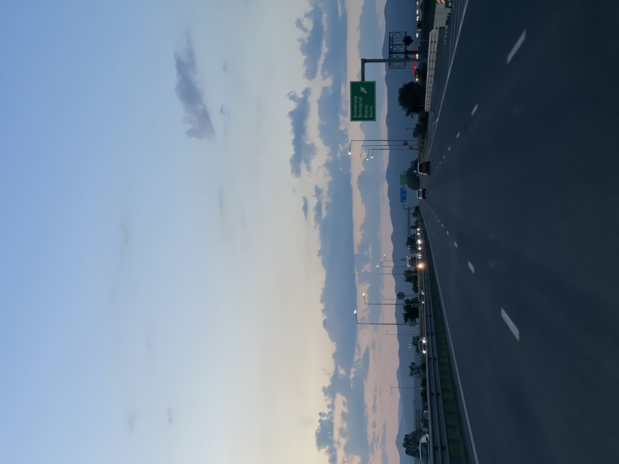 The image shows a highway in Krivina, Bulgaria. The highway stretches into the distance under a sky with scattered clouds. A green directional sign indicates routes to "Ботевград", "Витевград", and "Варна". Streetlights line the road, illuminating the scene. Cars are visible on the highway. Mountains are in the background. The image is oriented sideways.