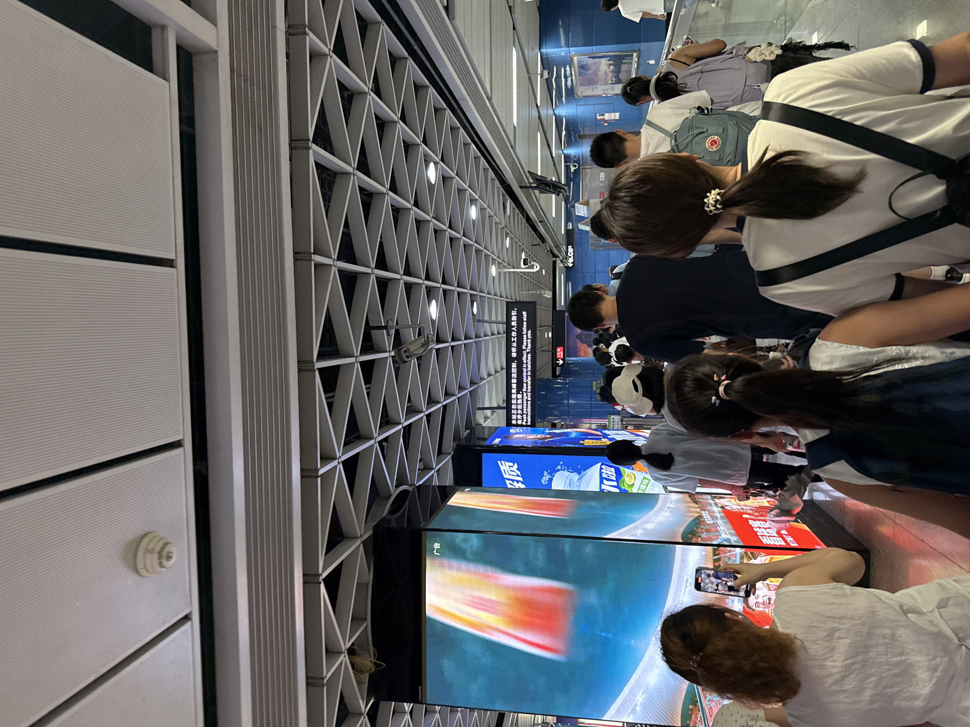The image depicts an indoor scene in Tianhe District, China, likely a subway station or an underground walkway, given the polished floor, large digital screens displaying advertisements, and the presence of multiple people walking in a single direction. The lighting appears to be artificial, with overhead fluorescent lights and illuminated screens.
In the foreground, several individuals are seen from behind. On the left, a woman with reddish-brown hair tied in a ponytail is holding up a smartphone, seemingly taking a picture or recording a video of one of the large screens. She is wearing a white sleeveless top. To her right, another person with dark hair pulled into a ponytail is wearing a white t-shirt with black straps from a backpack visible. Further back, a group of people are walking along a pathway. Some are carrying backpacks, including one with a blue backpack featuring a red logo.
The environment includes a modern, utilitarian ceiling structure made of metal grids and panels. Several large, vertically oriented digital screens are visible, showing vibrant advertisements with images and Chinese text. One screen on the left displays a close-up of what appears to be a sports event or a performance, with bright red and yellow colors. Another screen to its right shows a bottle of beverage and more Chinese text.
The overall atmosphere suggests a busy public space during the day, with people commuting or moving through the area. The focus of the image is on the people and their interaction with the advertising displays within this contemporary urban environment.