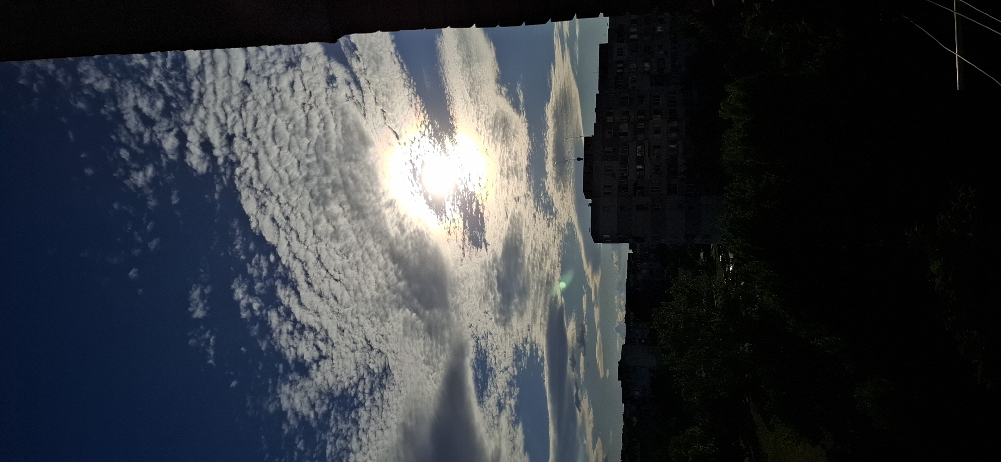 The image captures a sky filled with a textured pattern of altocumulus clouds, illuminated by the sun. The sun is partially obscured by clouds, creating a bright halo effect with subtle iridescent colors, a phenomenon known as a cloud iridescence or irisation. Below the sky, the silhouette of buildings and trees is visible against the bright sky. The buildings appear to be apartment blocks, characteristic of urban residential areas. The overall scene suggests an outdoor view, possibly from a balcony or window, overlooking a cityscape with a prominent sky. The time of day appears to be late afternoon or early evening, given the angle of the sun and the quality of light. The foreground is darkened, obscuring details of any immediate surroundings. This view is consistent with a residential neighborhood in Sofia, Bulgaria, where similar apartment blocks and tree-lined streets are common. There are no people or discernible actions depicted in the image.