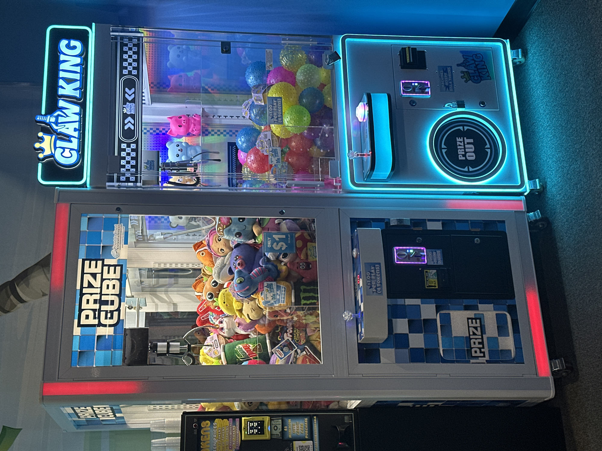 The image shows two arcade claw machines, side-by-side, in what appears to be an entertainment venue or arcade. The machine on the left is labeled "PRIZE CUBE" and is filled with plush toys. The machine on the right is labeled "CLAW KING" and contains colorful bouncy balls. Both machines have illuminated signage and colorful LED lighting. A token dispenser and payment area are visible on the left machine, with a credit card reader. The "CLAW KING" machine has a "PRIZE OUT" graphic and a token/bill acceptor. The machines are on casters, suggesting they are movable. The floor appears to be carpeted. The background is dimly lit, with a suggestion of a tropical-themed mural or decoration above the "PRIZE CUBE" machine. There are no people visible in the image. The setting does not provide specific clues to its location in Buffalo Grove, United States of America, beyond being an indoor arcade-style establishment.