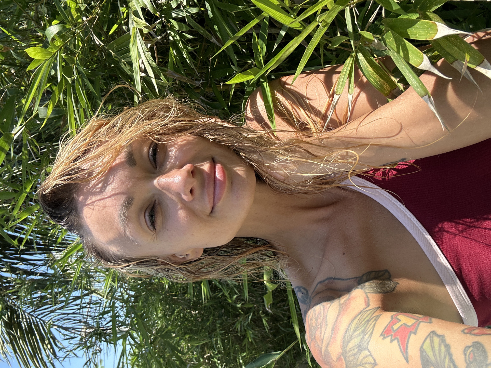 A woman with blonde hair, wet from the water, smiles at the camera. She is wearing a maroon top with a white trim. Her left arm is tattooed with various designs, including what appears to be a lightning bolt and flowers. She is surrounded by lush green foliage, possibly bamboo, and the sun shines brightly, casting shadows on her face and body. The scene suggests a tropical outdoor setting, potentially in Mũi Né, Vietnam, known for its beaches and coastal scenery.