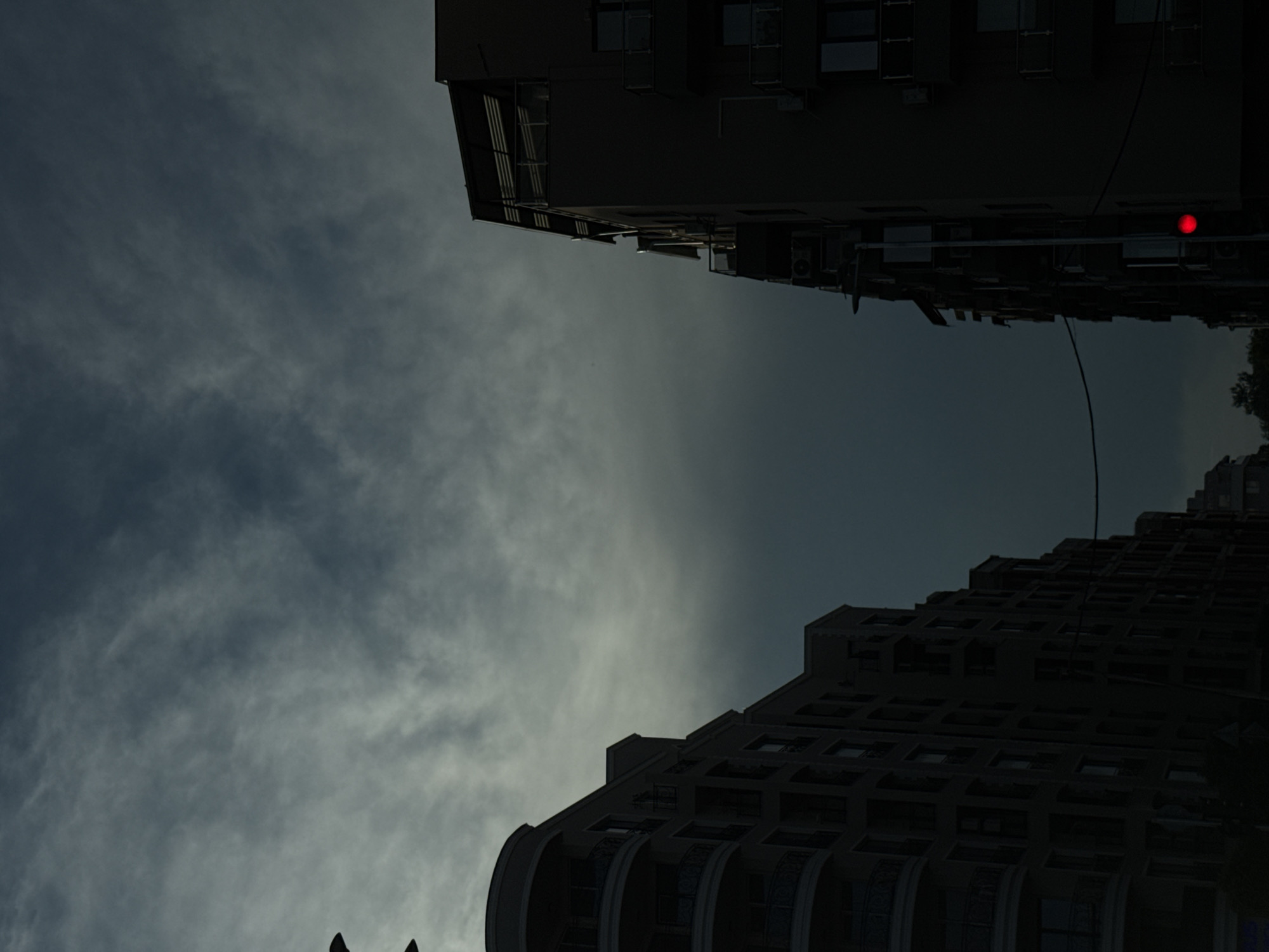 The image shows a view of two apartment buildings in Plovdiv, Bulgaria, under a cloudy sky. The buildings are dark and silhouetted, with balconies visible on the left building. On the right building, a red traffic light is visible. The sky is a deep gray with streaks of lighter clouds. The overall impression is one of a moody, overcast day in an urban setting.