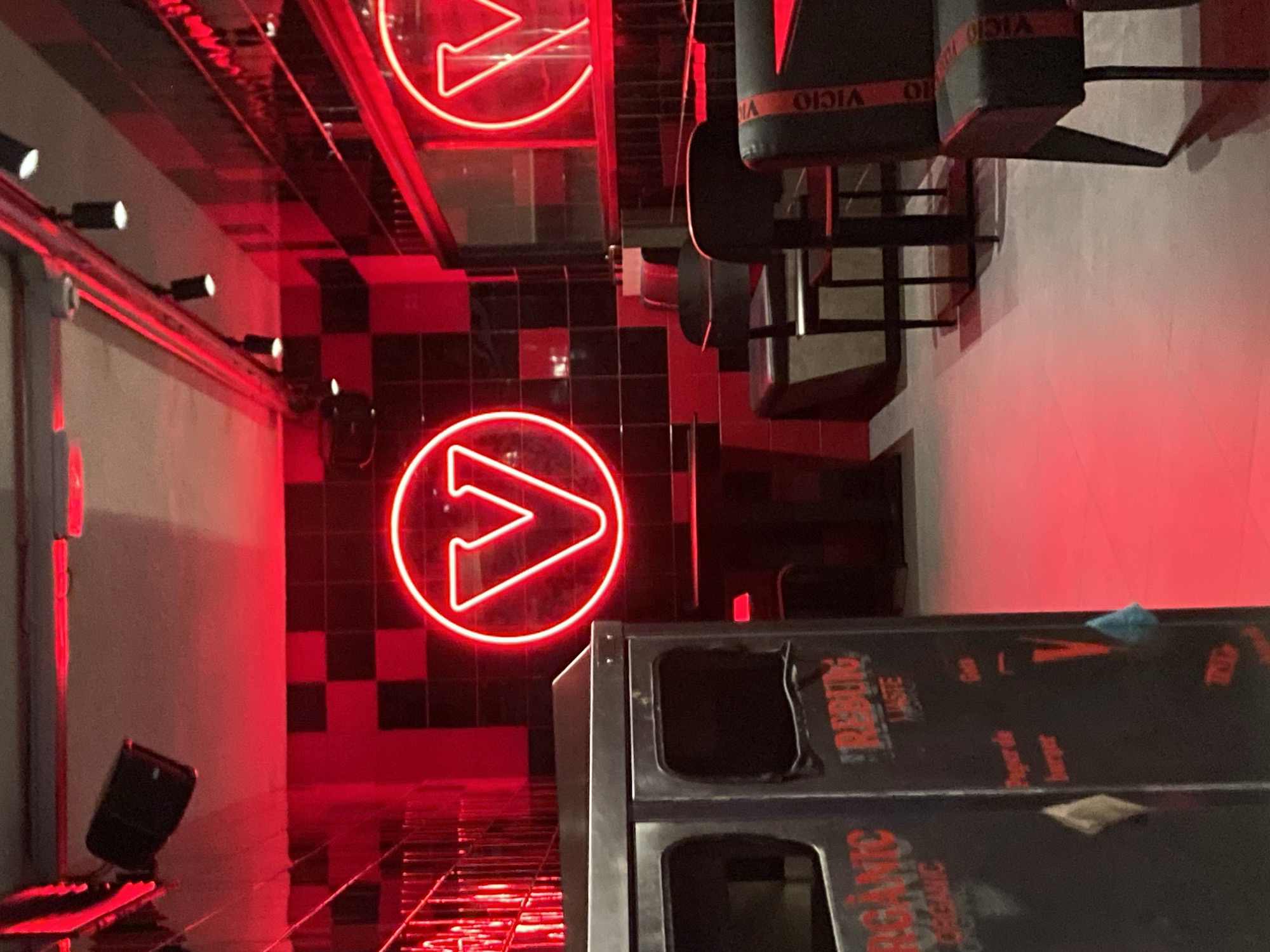 The image depicts the interior of a dimly lit restaurant with a striking red aesthetic. The walls are adorned with a black and red checkerboard pattern, and red neon lights cast a vibrant glow throughout the space. A prominent neon sign in the shape of a circle with a stylized 'V' inside illuminates the back wall. Several black tables and chairs are arranged in the dining area, some with red trim that reads "VICIO." A dark grey trash receptacle is visible in the foreground, with text that appears to be in French, indicating it is part of a "VICTOIRE ORGANIC" system, likely for waste disposal. The overall impression is of a modern, edgy dining establishment, possibly a fast-casual restaurant or a bar. Given the branding and apparent waste disposal system, this establishment could be related to a food service chain.
