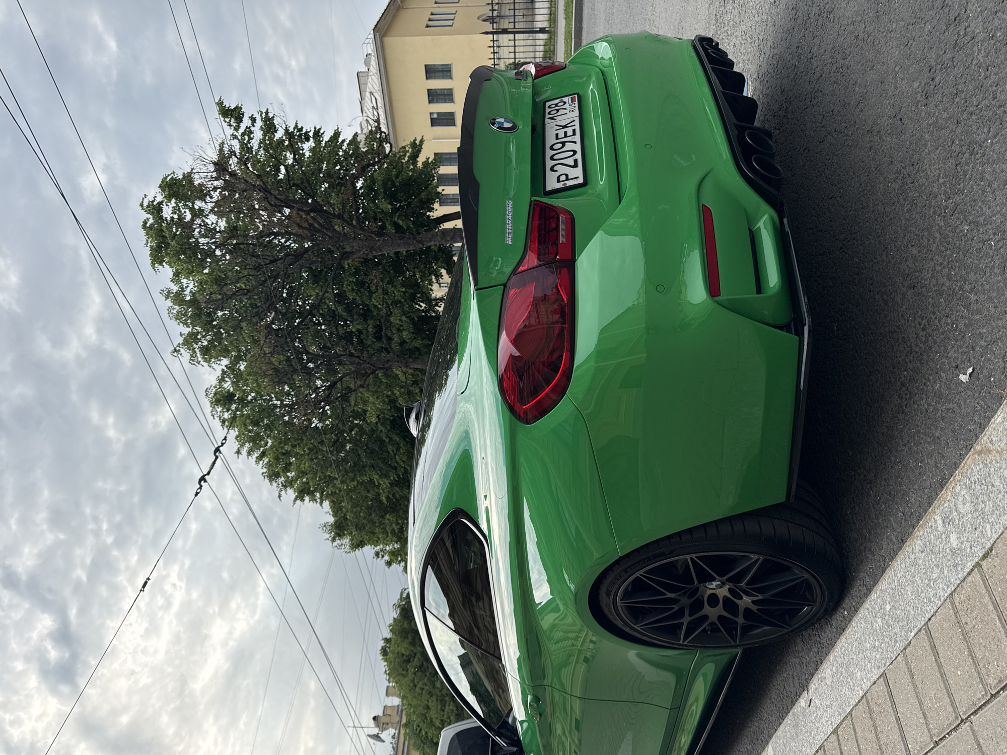 An eye-level medium shot shows the rear passenger side of a bright green BMW M4 coupe parked on a street in Saint Petersburg, Russia. The car is positioned slightly angled towards the right, with its rear facing the viewer. The vehicle has a spoiler on the trunk, quad exhaust pipes, and black M-style wheels. A license plate reading "P209EK 198" is visible on the rear bumper.
The car is parked on asphalt next to a sidewalk made of gray paving stones and a granite curb. In the background, a yellow building with multiple windows and a dark fence are visible, along with lush green trees and a cloudy sky. Overhead, power lines crisscross the sky. The overall scene suggests an urban environment on an overcast day. No people are visible in the image.