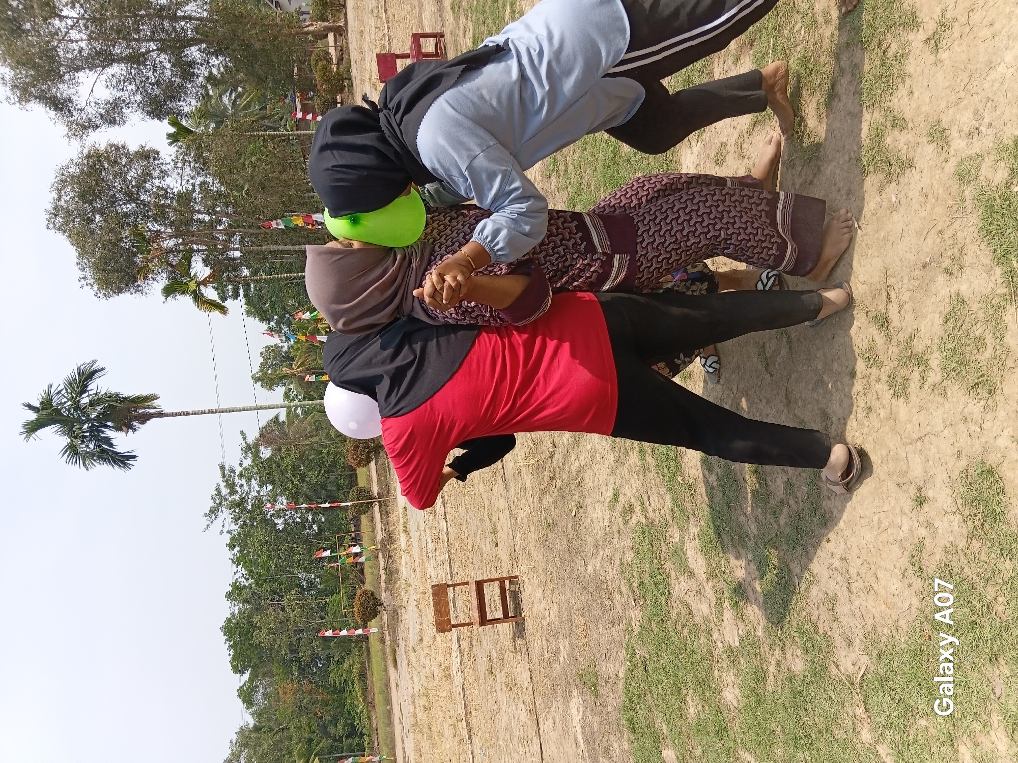 Three individuals, two women and one partially visible person, are engaged in a game outdoors in what appears to be a field or park area. The two visible women are wearing hijabs, one in brown and the other in black. The woman in the brown hijab is dressed in a patterned maroon and white dress, while the woman in the black hijab wears a light blue long-sleeved shirt and black pants with white stripes. The third individual, facing away from the camera, is wearing a red long-sleeved shirt and black pants, with a white balloon obscuring their face. The two women are holding a green balloon between their faces, appearing to be trying to pop it or keep it inflated. They are also holding hands. A simple wooden chair is positioned in the background to the left. The ground is a mixture of dry dirt and sparse green grass. In the background, several trees, some tall and slender, others bushy, form a backdrop. Red and white Indonesian flags are visible on poles in the mid-ground. The sky is clear and bright, suggesting it is daytime. The scene is likely part of a community event or recreational activity in Margo Rukun, Indonesia, possibly related to a celebration given the presence of the flags.