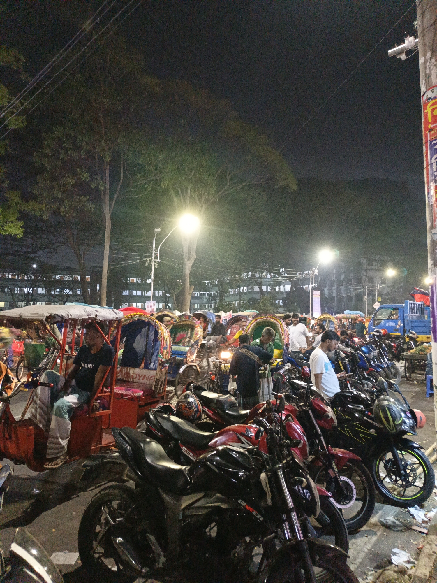 The image depicts a street scene in Dhaka, Bangladesh, at night. In the foreground, there are rows of parked motorcycles of various makes and models. Several cycle rickshaws, also known as "rickshaws," are visible, some occupied by drivers. In the middle ground, people are standing and talking in groups. In the background, a multi-story building with illuminated windows is visible. Streetlights provide illumination. A utility pole with visible wiring and advertisements stands to the right. Debris is scattered on the ground.