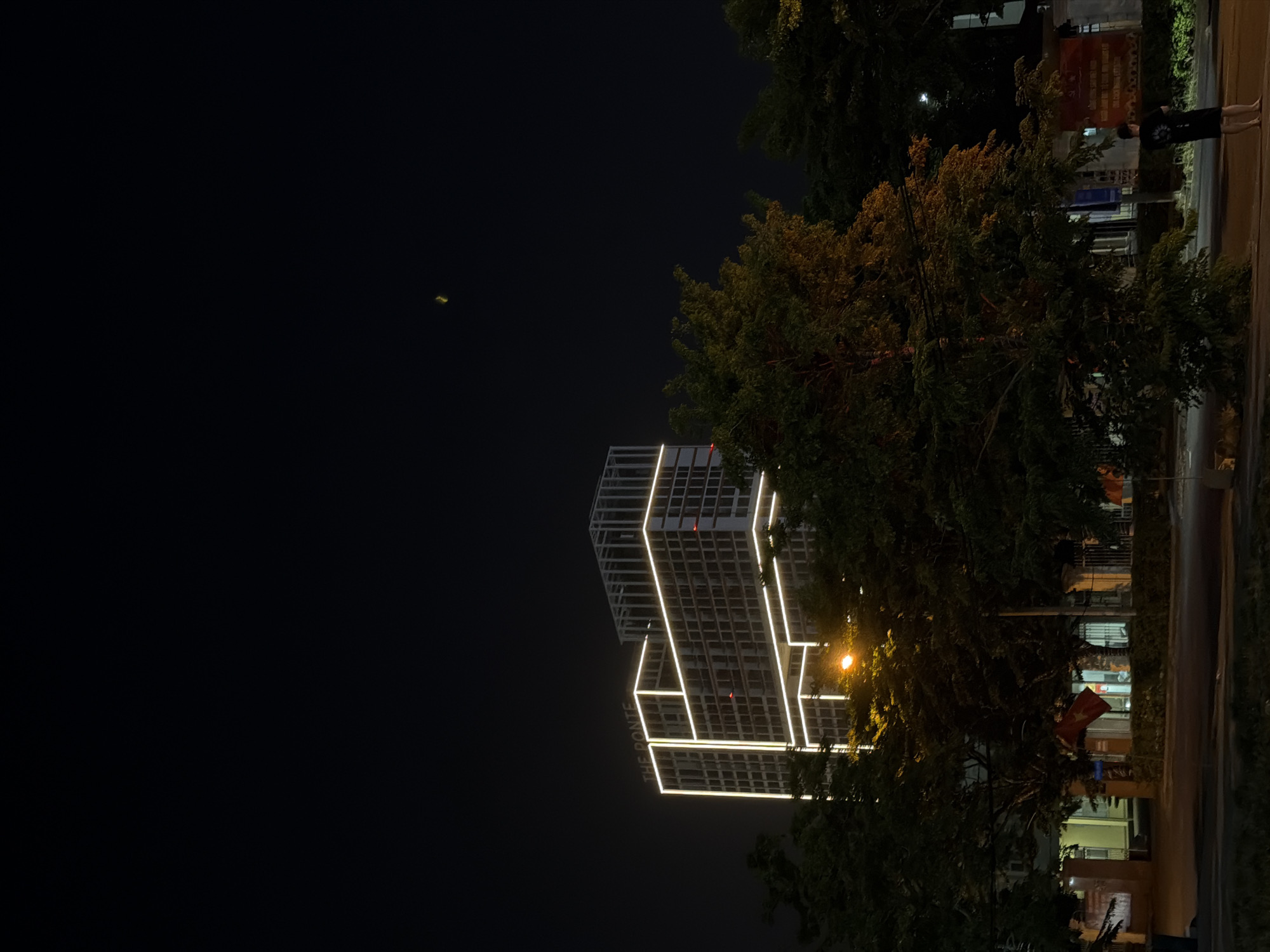 At night, a modern building with illuminated white lines outlining its structure stands adjacent to a tree with dense foliage. A street borders the edge of the frame with a single person standing on it. This building appears to be "The Ponte" hotel, located in Đà Nẵng, Vietnam.