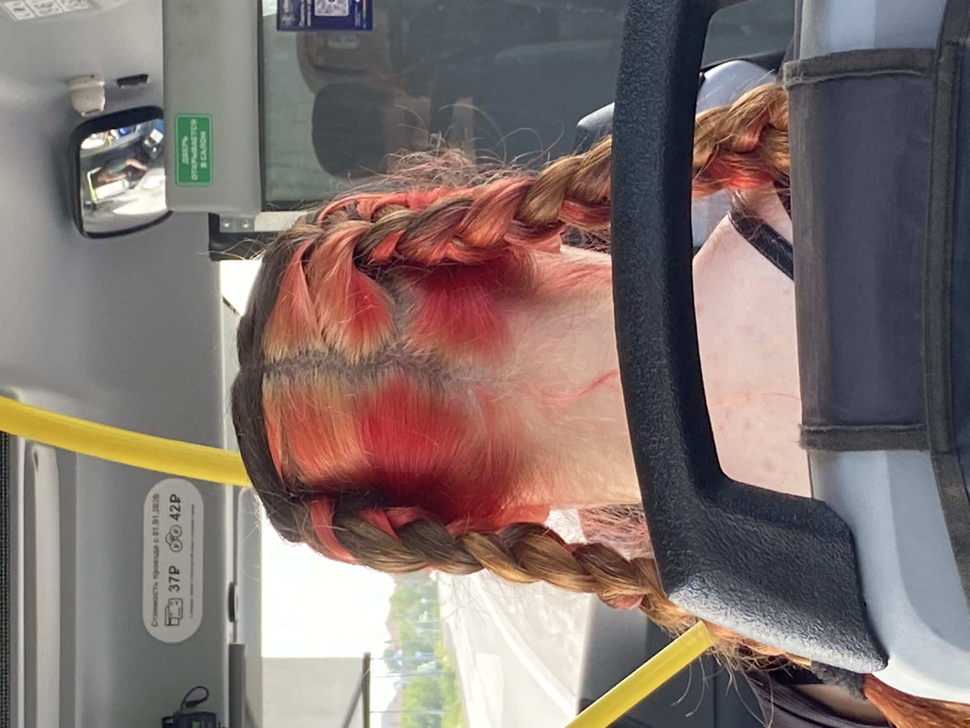 The image shows the back of a person's head inside a bus, likely in the city of Tyumen, Russia, based on the presence of Cyrillic text on signs. The person has long hair styled in two braids, with the hair dyed in a gradient of reddish-orange and brown tones. The hair is pulled back from the face and tied into braids that extend down the back. Some of the hair appears to be naturally brown, while other sections have been dyed a vibrant red. The background of the image shows the interior of a bus, with a yellow pole visible to the left and a black handrail in the foreground. A mirror is also visible, reflecting a person's face and shoulders. Signs with Cyrillic text are present on the bus ceiling, indicating information about fares and door operation. The overall lighting suggests daytime.