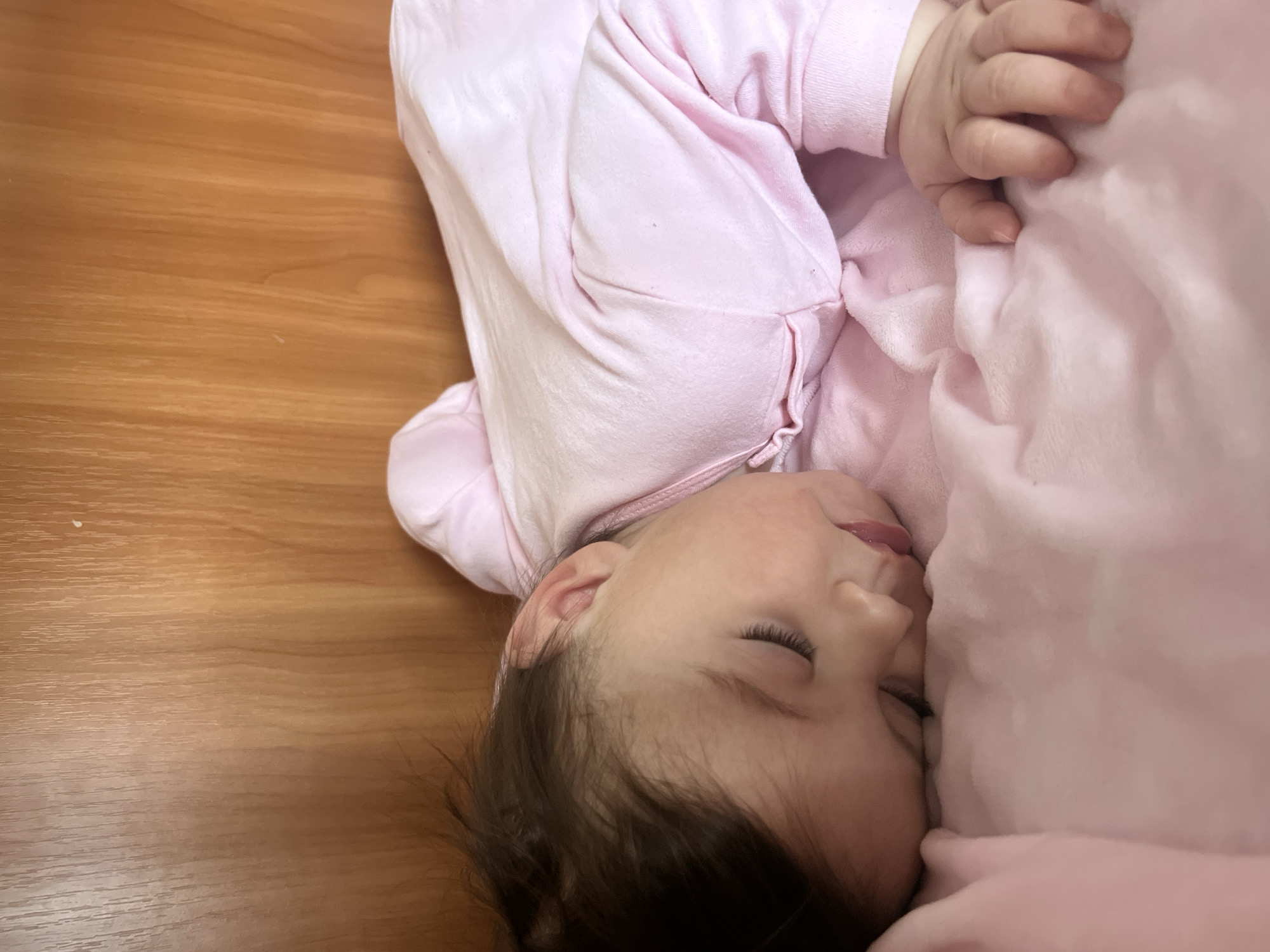 A young child, possibly a baby or toddler, is lying asleep. The child is dressed in a light pink outfit and is nestled against a light pink blanket or pillow. The child's dark hair is visible against their skin. They appear to be lying on a wooden floor or surface. There is nothing in the image to indicate the location is Elena, Bulgaria.