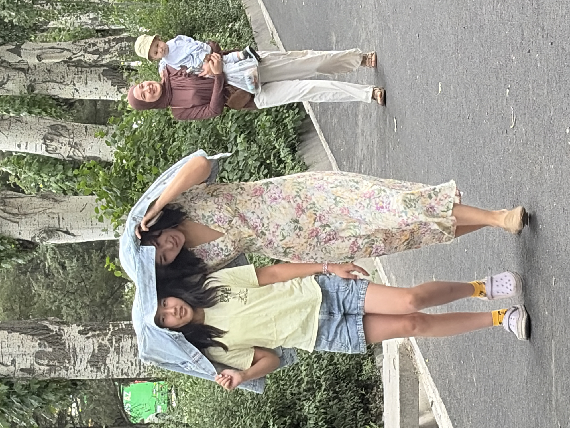 In an outdoor, urban setting, three individuals are walking on a paved path lined with trees and greenery. In the foreground, a young woman and a girl walk side-by-side. The girl, on the left, wears a light yellow t-shirt, denim shorts, and white and purple clogs with yellow socks. She holds a light blue denim jacket above her head, sharing it as a makeshift umbrella with the woman next to her. The woman, on the right, wears a floral-print dress. Behind them, a woman in a maroon hijab and brown top, white pants, and brown sandals carries a baby in a blue outfit and a pale yellow hat. The baby is held in her left arm. The path is a dark asphalt with scattered debris. In the background, lush green foliage and the patterned trunks of trees are visible. A bright green vehicle is partially visible to the far left.