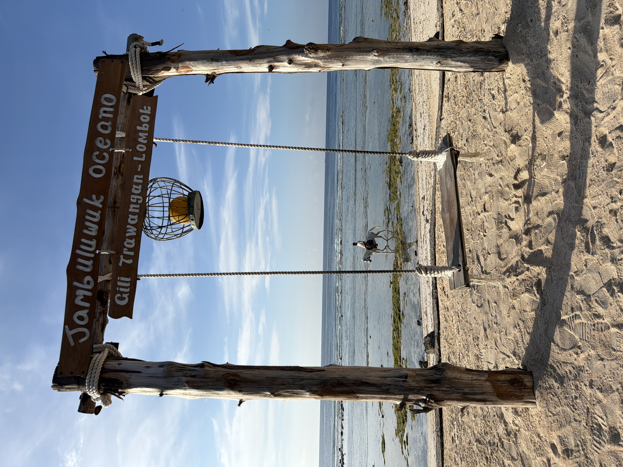 A wooden swing hangs from a weathered timber frame on a sandy beach. The swing set has a sign overhead that reads "Jambululuk Oceano" and "Gili Trawangan-Lombok". A wire basket with a yellow orb hangs from the sign. In the background, the ocean stretches out under a blue sky with scattered clouds. A person is riding a white horse in the shallow water near the shore. The sand is imprinted with footprints and tire tracks.