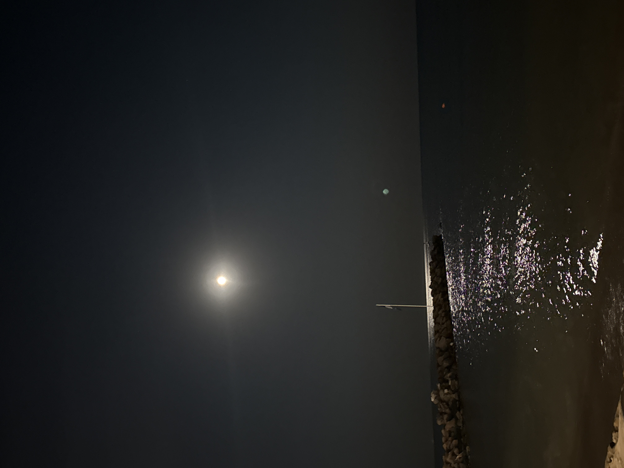 The image depicts a nighttime scene on the coast of Sveti Vlas, Bulgaria. A full moon is prominently visible in the dark sky, casting a bright glow. Below the moon, a pier made of large rocks extends into the calm, dark sea. A white flagpole stands at the end of the pier. The moonlight reflects off the water, creating a shimmering path. The foreground shows a dark beach area. The overall atmosphere is serene and tranquil.