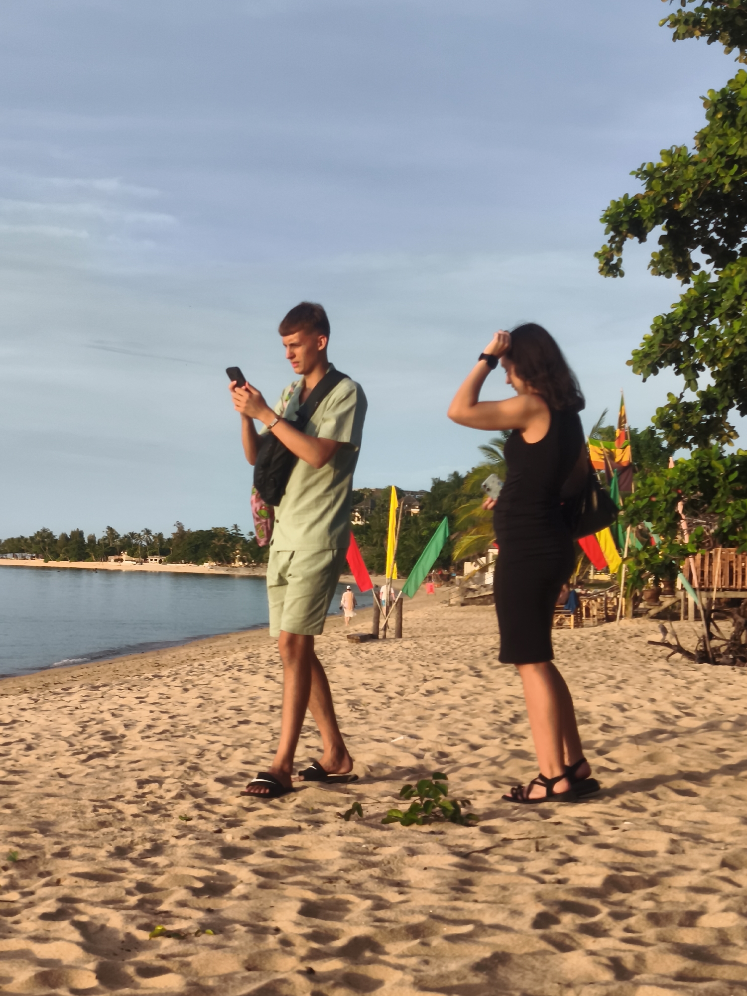 The image depicts a beach scene in Baan Bo Phut, Thailand. Two individuals are visible on the sandy shore. In the foreground, a young man stands facing left, holding a smartphone in his hands. He is wearing a light green short-sleeved shirt and matching shorts, with black flip-flops on his feet. A black bag is slung across his chest. To his right and slightly behind him, a woman stands facing left with her right hand raised to her head. She is wearing a black sleeveless dress and black sandals. She also has a black bag slung over her shoulder. The beach is characterized by soft sand with some sparse green foliage. In the background, the calm sea is visible, with a line of palm trees and buildings along the coastline. Several colorful flags on poles are also present on the beach, adding to the tropical ambiance. The sky is a pale blue with some wispy clouds. The lighting suggests it is late afternoon or early evening.