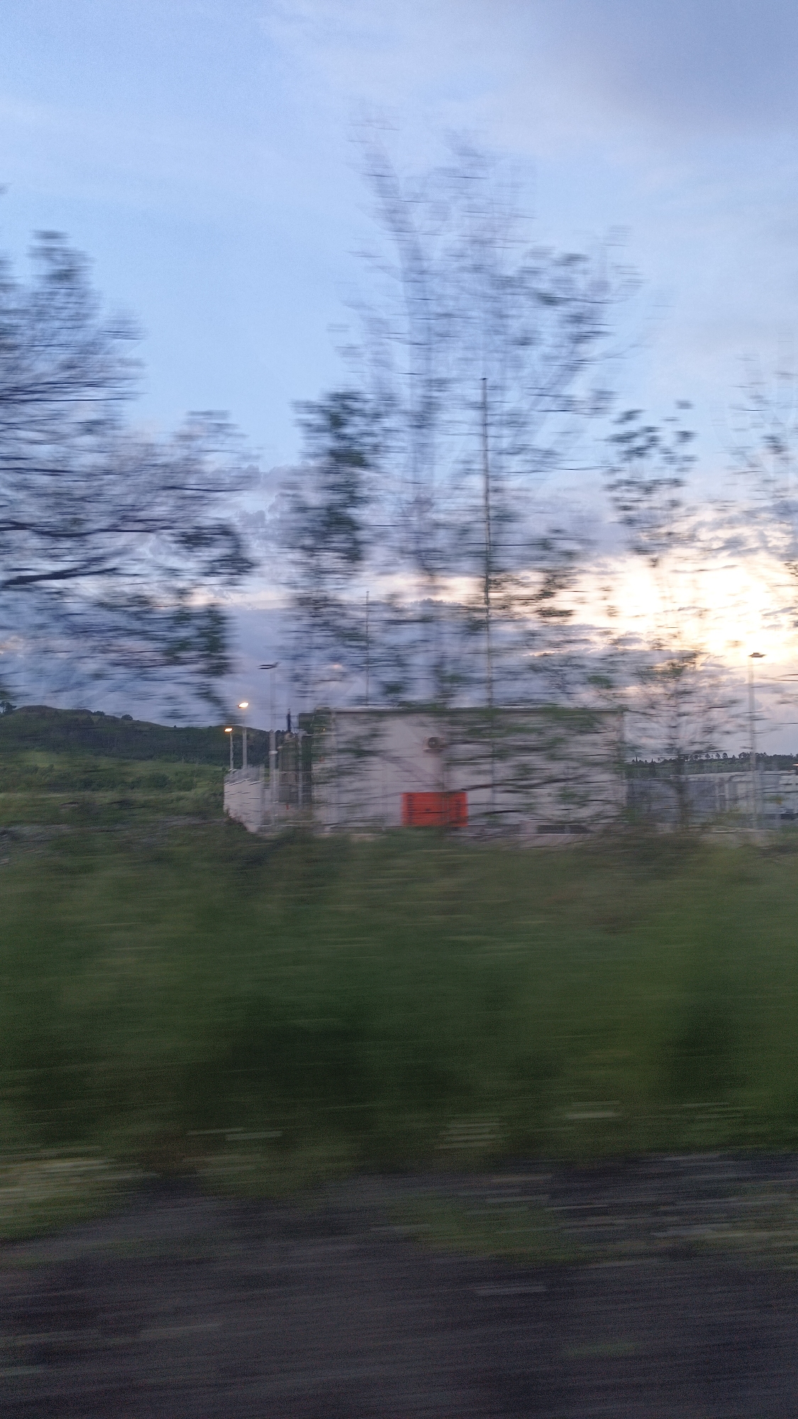 The image depicts a blurred landscape, likely taken from a moving vehicle. In the foreground, there is blurred foliage and a portion of a dark surface, possibly a road or ground. In the middle ground, there is a light-colored building with an orange or red element, potentially a door or panel. There is also what appears to be a radio tower or some other type of infrastructure extending vertically. In the background, there are rolling hills and a sky with a mix of blue and white clouds, suggesting either dawn or dusk. Given the presence of the radio tower and the landscape features, it is plausible the location is Burgas, Bulgaria, but the blurriness of the image prevents a definitive confirmation. The scene lacks any discernible people or clear actions.