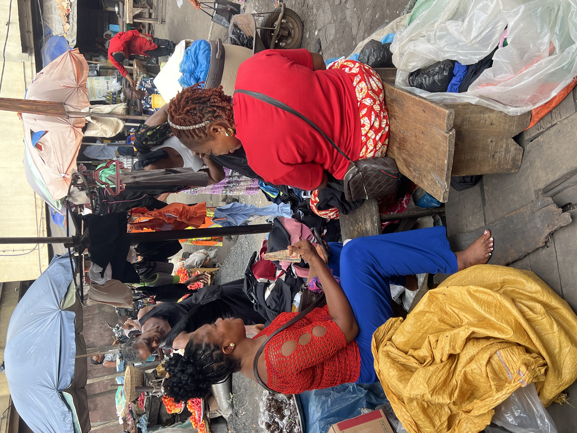 This image captures a vibrant open-air market scene in Nkpor, Nigeria. The setting is bustling, with various goods displayed around multiple individuals, mostly women.
In the foreground, two women are prominent. One woman, with dreadlocks and a white beaded headband, wears a bright red top and a red-and-white patterned skirt. She is seated on a wooden bench, facing away from the camera, with a dark shoulder bag slung across her. Beside her, another woman in a red, intricately patterned mesh-like top and vibrant blue pants sits on a lower surface. She has braided hair and is holding Nigerian Naira currency, specifically a 500 Naira note, suggesting a transaction or the intent to purchase. Both women wear gold hoop earrings. The woman in blue pants is barefoot.
Around them, various merchandise is visible. Clothes are hung on racks and piled on surfaces, including what appears to be a wooden stall or crate. Plastic bags, some clear and others blue or black, contain unseen goods. A large yellow woven sack rests on the ground next to the woman in blue pants. In the background, multiple umbrellas, one light blue and another beige, provide shade from the bright daylight. Other market-goers and vendors are partially visible, including an older man with glasses seated under an umbrella. Trays holding what might be food items or small packaged goods are also present. The ground is a mix of concrete and dirt, typical of an informal market. The scene conveys the busy atmosphere of daily commerce.
Visible text is limited; a partially obscured package in the background reads "MAY YOUR WORLD... SHOW BIG" (possibly "SHOW BIZ").