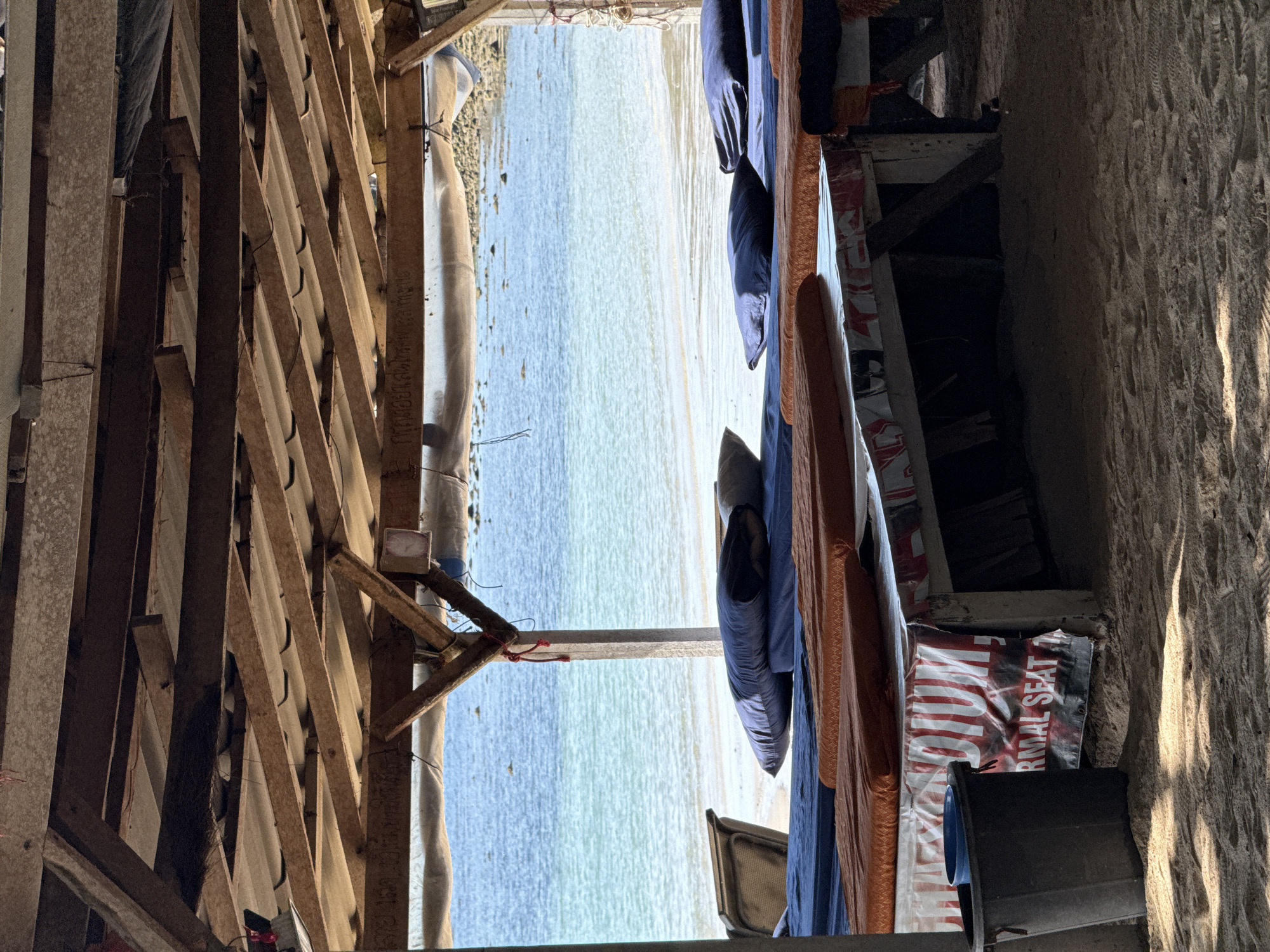 The image depicts an outdoor seating area on a sandy beach, likely in Baan Chaweng Noi, Thailand, overlooking the ocean. The foreground is dominated by several low-lying tables covered with blue and orange cushions. A dark blue bucket sits to the left of the tables. The tables themselves are covered with a banner that has the word "DIUM" and "RMAL SEAT 1" visible. Above the seating area, a wooden roof structure is visible, constructed from various wooden planks and beams, with corrugated material underneath. The background reveals a calm, turquoise ocean with a sandy beach and some rocks or coral formations visible in the water. The lighting suggests it is daytime. There are no people or animals visible in the scene.