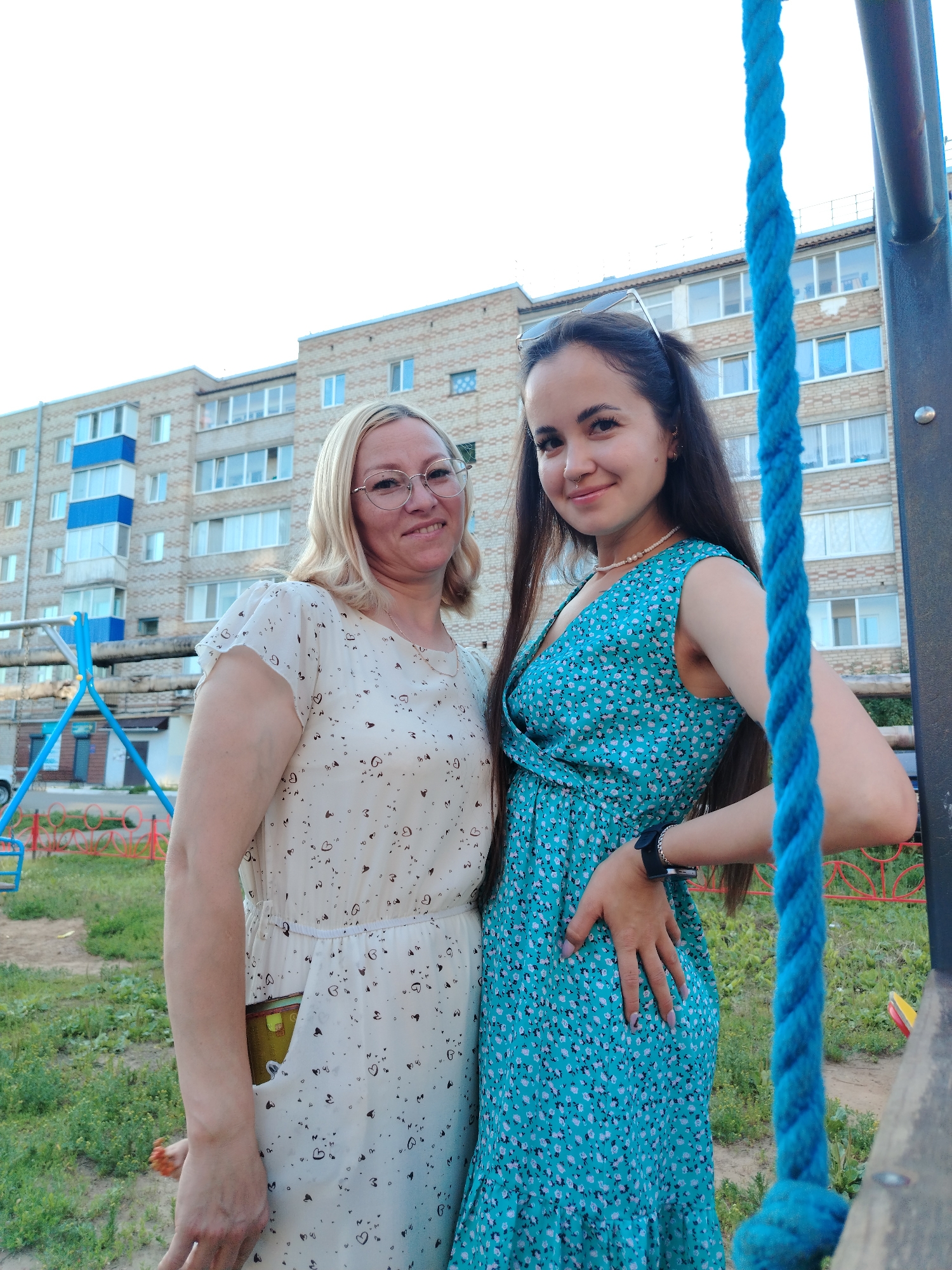 In Yanaul, Russia, two individuals are captured in a full-frame, low-angle outdoor photograph, standing against the backdrop of a beige apartment building. The building, featuring light-colored brickwork with blue and white accents on its balconies, occupies the upper half of the image. A playground structure, with blue and red elements, is visible to the left.
On the left, a woman with blonde hair, wearing glasses and a light-colored dress adorned with a subtle heart pattern, stands with her hands at her sides. A yellow object protrudes from her dress pocket. To her right, a younger woman with long, dark hair, sporting a turquoise floral dress, rests her hand on her hip. She wears a watch on her left wrist and has sunglasses perched on her head. A thick, braided blue rope hangs prominently in the foreground, partially obscuring the younger woman. The ground is covered in patchy green grass. The sky above is a light, overcast white.