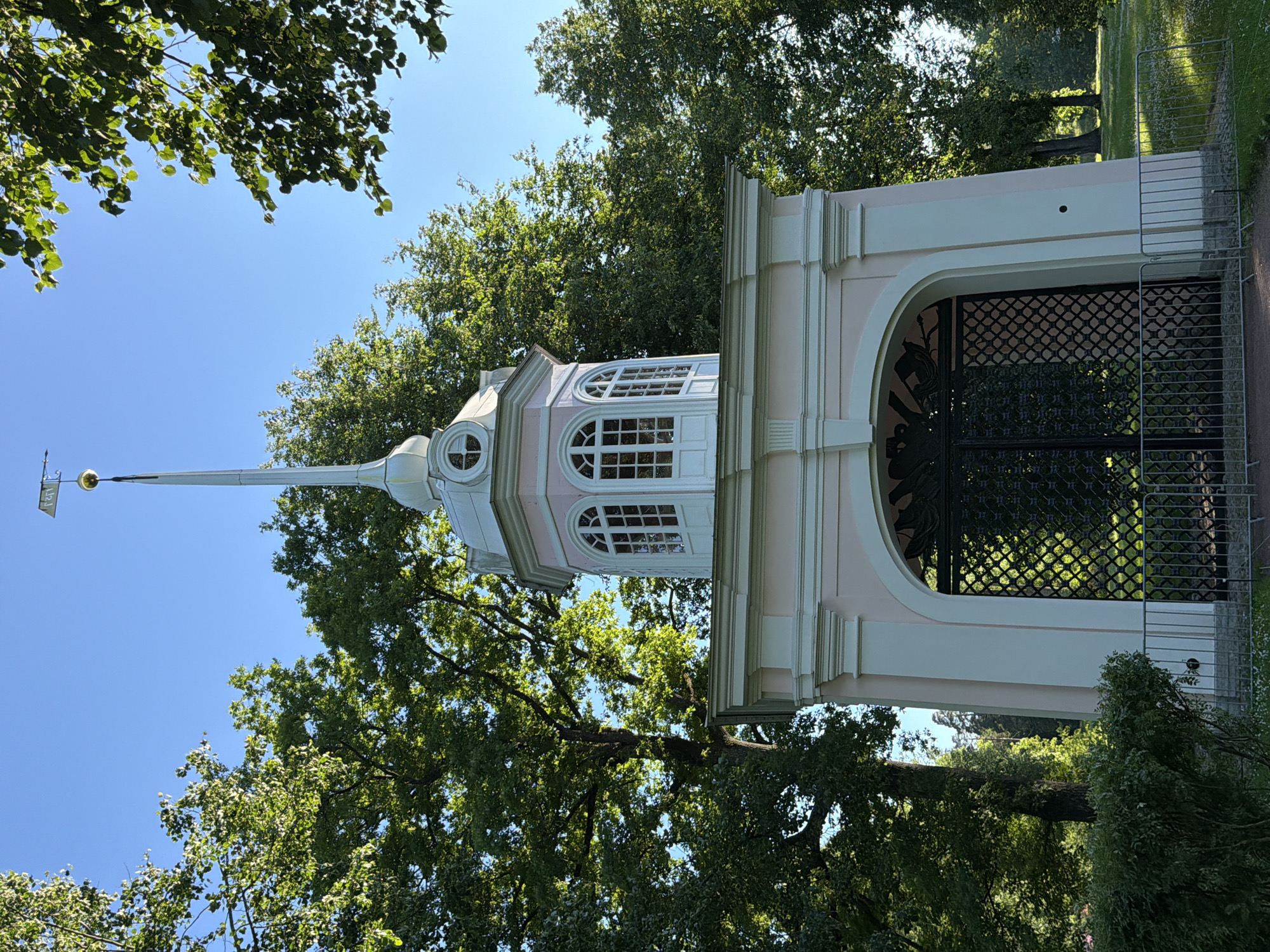 The image depicts a pale pink and white ornate pavilion topped with a spire and a weathervane. The pavilion has an arched entryway with decorative black metal gates. Above the entryway, there are tall windows with arched tops. The structure is surrounded by lush green trees and a clear blue sky. In the foreground, there is a low metal fence and some greenery. The scene appears to be in a park or garden setting.