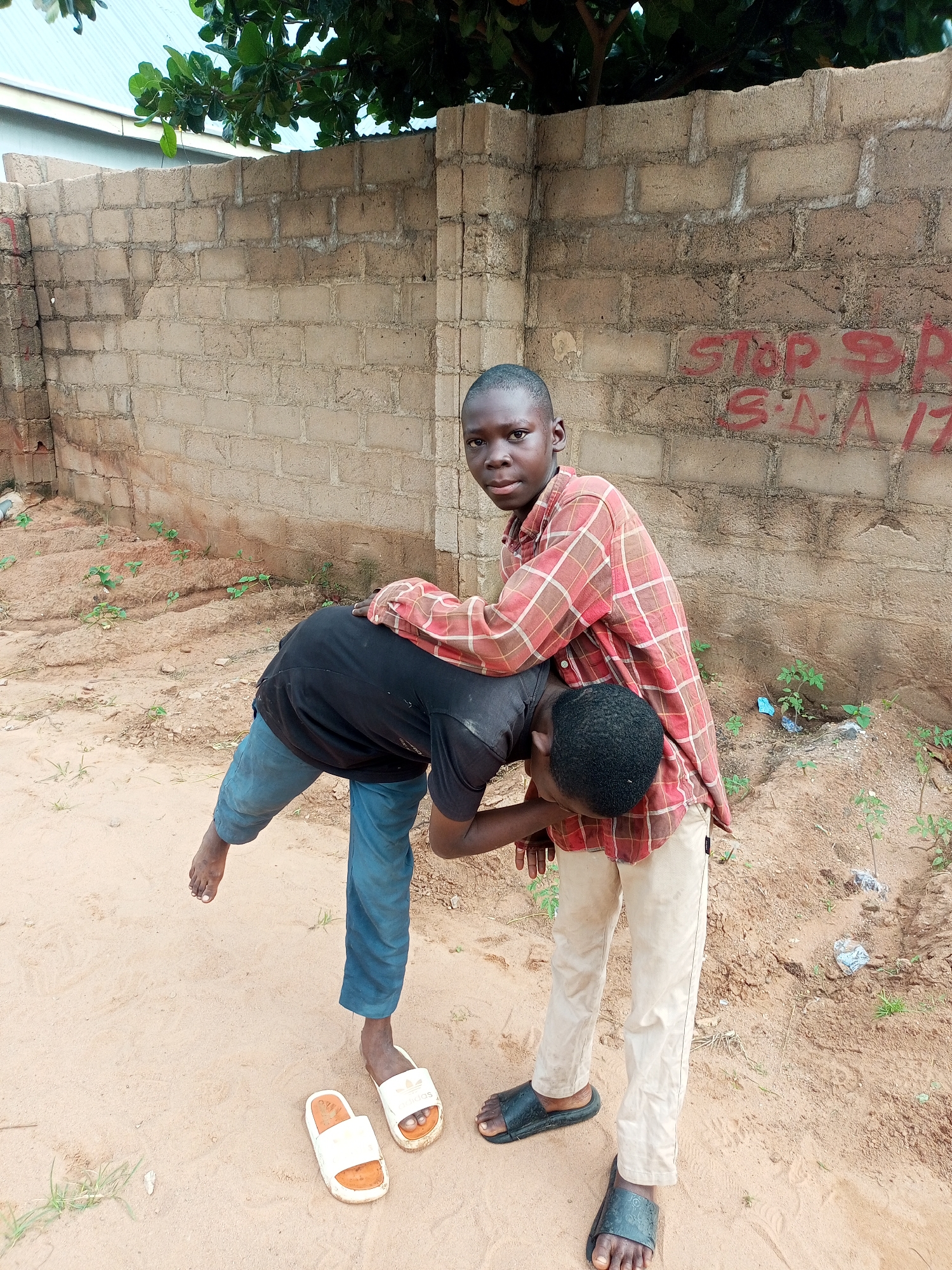 Two young boys are engaged in playful wrestling or a similar physical interaction outdoors. The boy on the right, wearing a red and brown plaid shirt and light-colored pants, has his arms around the neck and upper back of the boy on the left. The boy on the left, dressed in a black t-shirt and blue jeans, is bent over, with his left leg lifted off the ground. He is wearing white flip-flops, one of which has fallen off. The boy on the right is wearing dark slide sandals.
They are standing on sandy ground with a few patches of sparse vegetation. Behind them is a brick wall, some sections of which appear weathered or damaged. Red graffiti is visible on the wall, with the words "STOP SP" and "SA 17" clearly discernible. Above the wall, a tree with green leaves and a portion of a light-colored building or roof are visible. The lighting suggests it is daytime, possibly morning or afternoon, with no strong indications of extreme weather conditions. The mood appears to be one of youthful energy and play. The location is identified as Tempure, Nigeria.