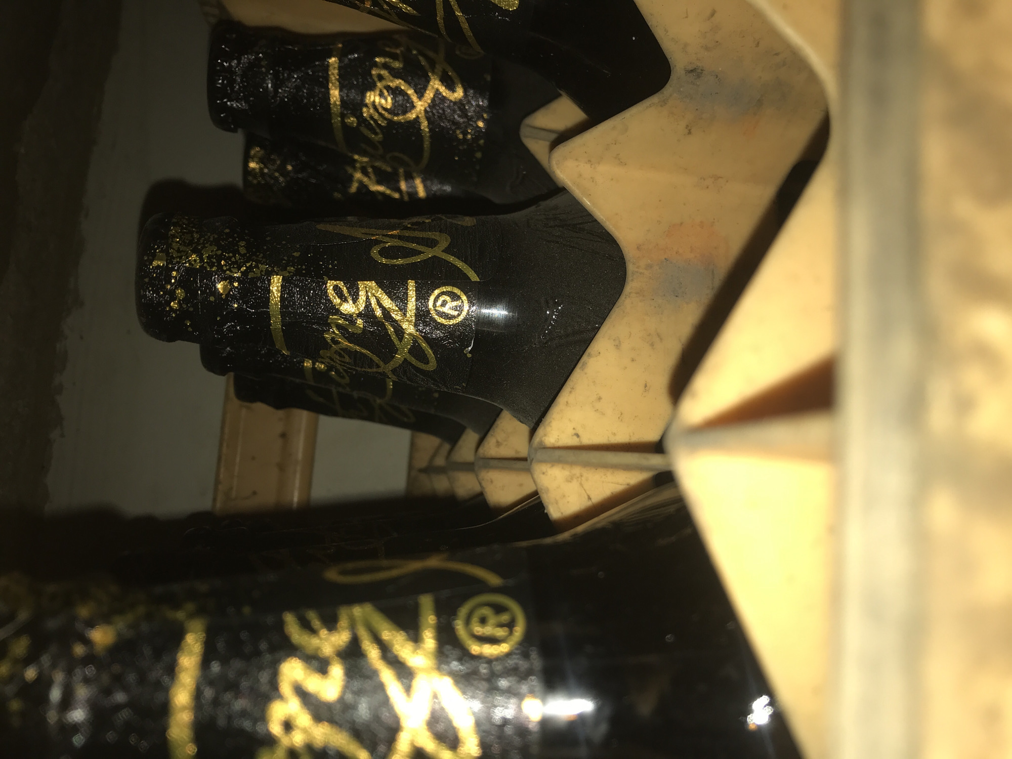 In a dimly lit room, possibly a storage area in New Stadium, Nigeria, a crate of dark beer bottles is visible. The bottles are black with metallic gold lettering and accents. The labels appear to have a cursive script and a registered trademark symbol of an "R" within a circle on at least one of them. The bottles are arranged in an orange plastic crate, with some appearing to be covered in dust or a light coating of something. The focus is on the upper portions of the bottles, with the crate and background out of focus. The overall impression is one of stacked beverage bottles in a dimly lit, possibly aged, environment.