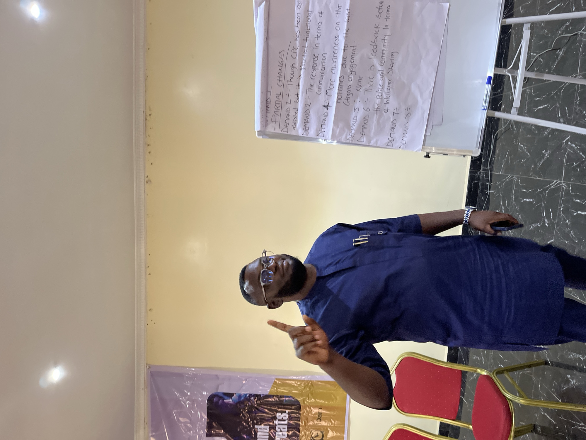A man in a blue shirt and glasses is standing indoors, pointing his index finger up. Behind him is a whiteboard with a list titled "DEMAND I - PARTIAL CHANGES" written in capital letters. The list is broken down into several points labeled "DEMAND 1" through "DEMAND 8," with brief descriptions of each. Red chairs and a banner with the word "eats" and "10ar" are also visible. The image was taken in the city of Gura Top, Nigeria.