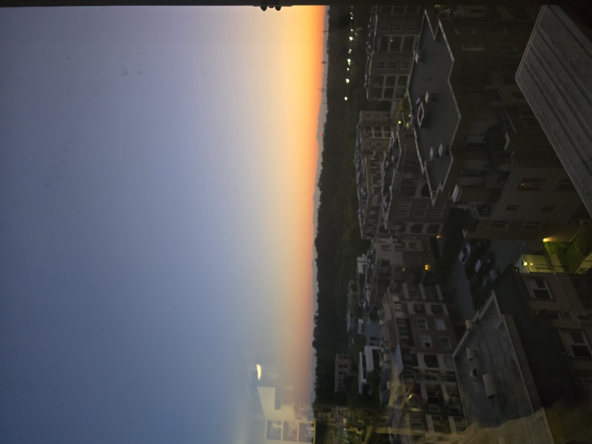 The image shows a high-angle view of a cityscape at dusk or dawn. The sky transitions from a dark blue to a bright orange along the horizon. A dense line of trees separates the sky from the urban landscape. Buildings of varying heights are densely packed together, with windows reflecting light. A tall, gray structure stands prominently on the right side of the frame. Streetlights are visible, adding to the ambiance of the scene. The specific location in the city of Unknown, Unknown is indistinguishable, but the scene suggests a densely populated urban area. There are no people visible in the image.