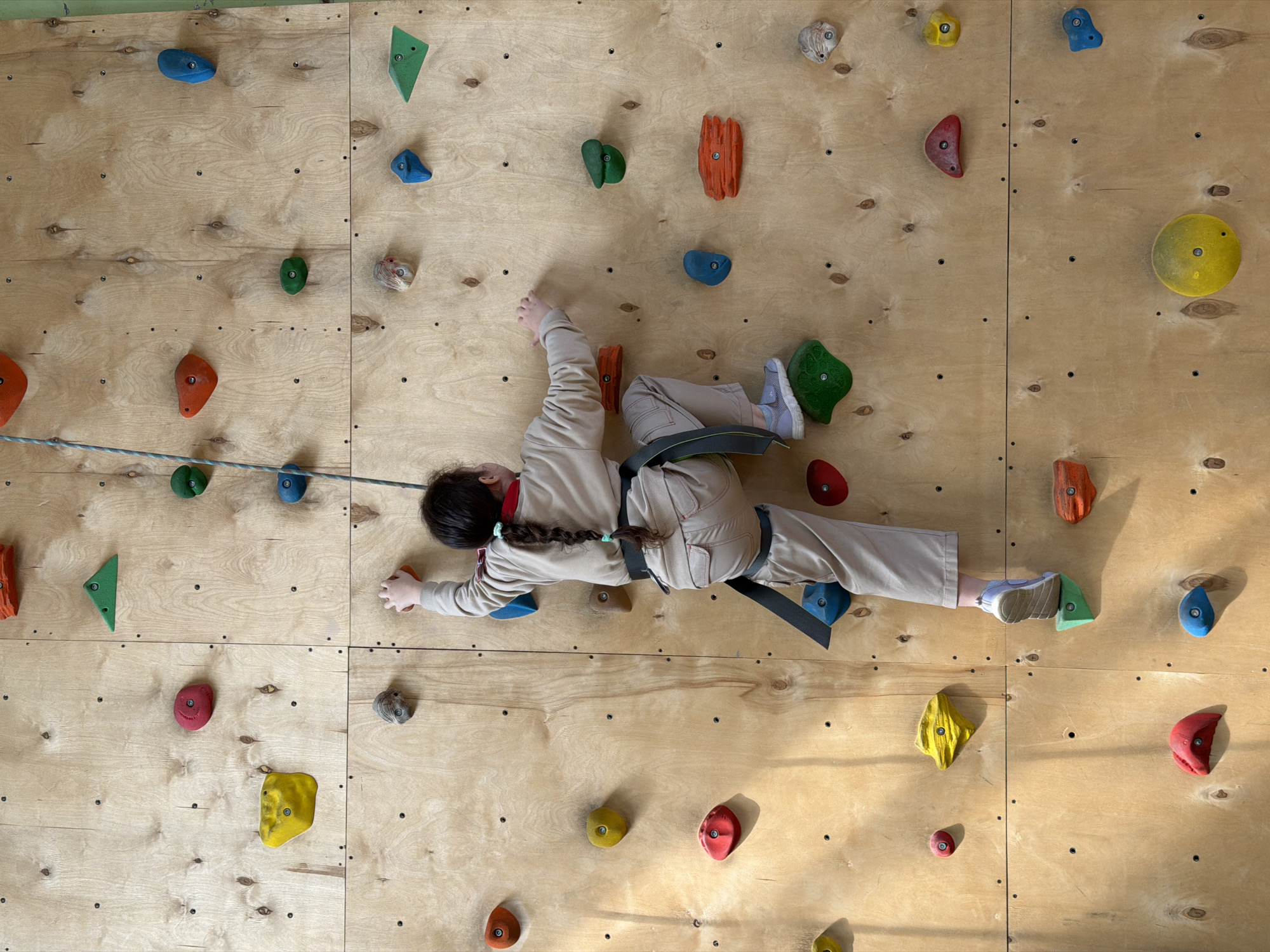 A child is shown rock climbing on an indoor climbing wall. The climbing wall is constructed from large plywood panels and features a variety of colorful climbing holds. The child is wearing beige pants, a beige top, a climbing harness, and athletic shoes. A rope extends from the harness. The location is Izhevsk, Russia, as stated.