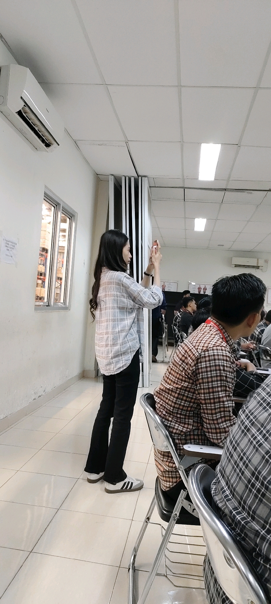 In a classroom setting in Nusa Indah, Indonesia, a woman with long, dark hair is standing on the left side of the frame, facing away from the camera. She is wearing a light-colored plaid shirt and black pants, with white sneakers. She holds a mobile phone in both hands, appearing to take a photo or record a video. In the background, several other individuals are seated at desks, also facing away from the camera. They are wearing various plaid shirts, suggesting a dress code or a shared affiliation. The room is brightly lit by overhead fluorescent lights and features a tiled floor and white walls. An air conditioning unit is mounted high on the wall to the left, and a window showing shelves of what appear to be stacked boxes is visible. Movable partitions are positioned behind the standing woman. The overall impression is that of a classroom or lecture hall during a presentation or event.