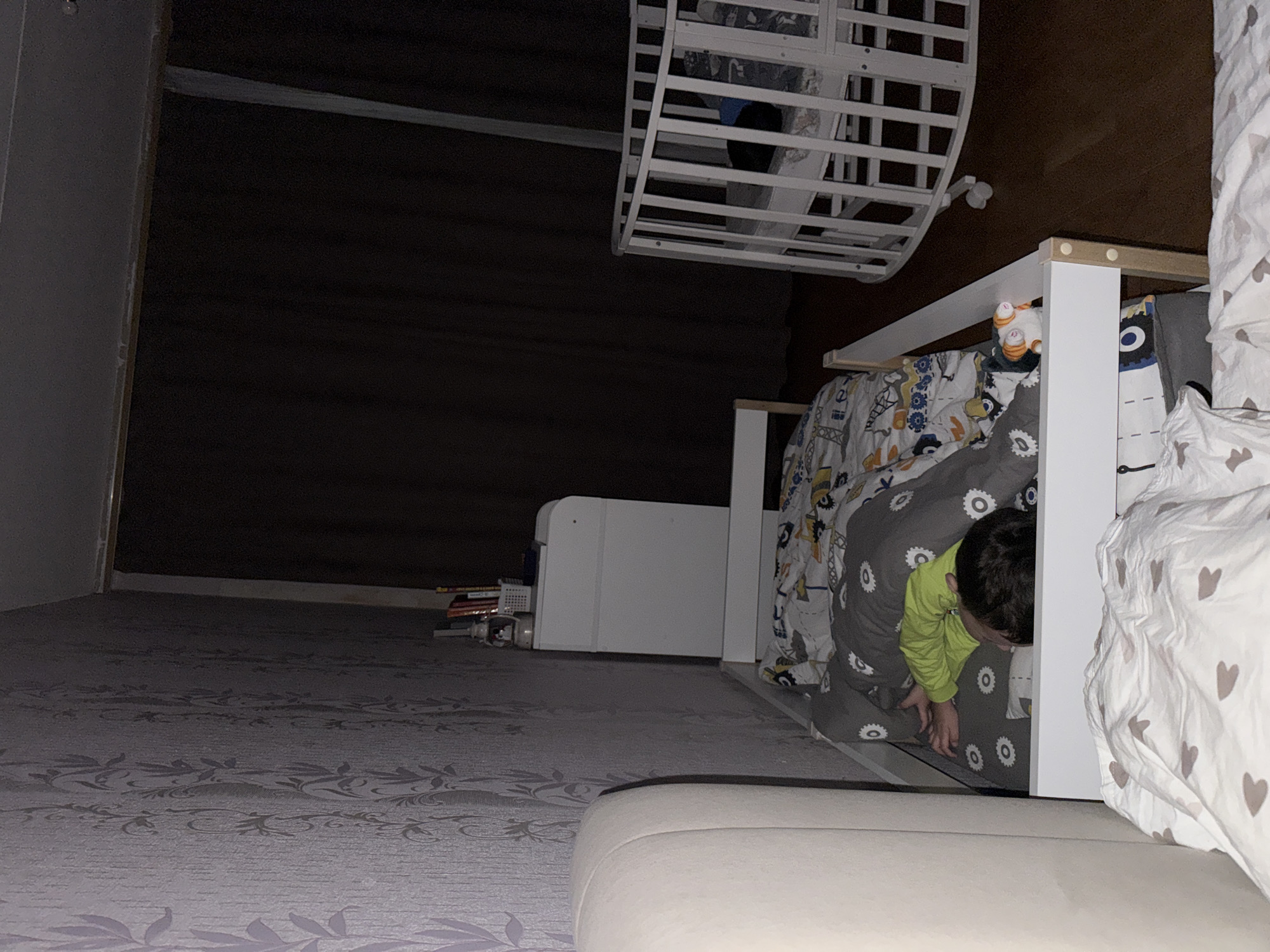 A dimly lit bedroom is captured in this photograph. The focal point is a child lying in a bed with a gray and white patterned comforter. The child is wearing a lime green long-sleeved shirt. The bed has a white frame. A second bed with white bars sits atop the first bed, presumably a bunk bed. A white storage unit is situated near the bed. On the floor near the storage unit, there are several books and miscellaneous items. The room's background features a dark wall or curtain, contributing to the overall dimness. The location is Chaikovsky, Russia.
