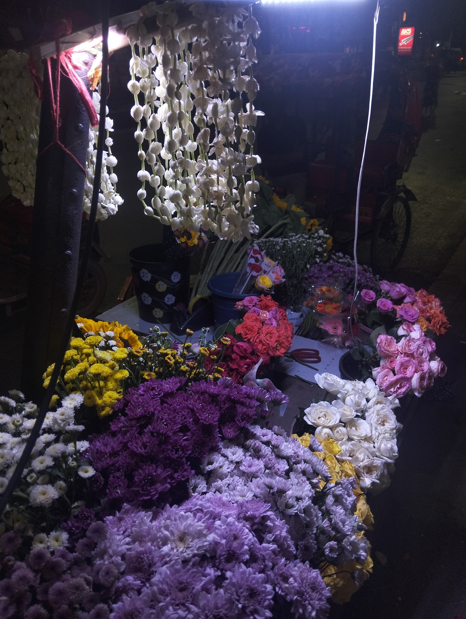 A flower stall in Dhaka, Bangladesh is brightly lit at night. Bunches of colorful flowers, including roses, chrysanthemums, and sunflowers, are displayed on a table. Strings of white jasmine garlands hang from the top of the stall. A bicycle is parked nearby. A sign with "AKS" on it is visible in the background.
