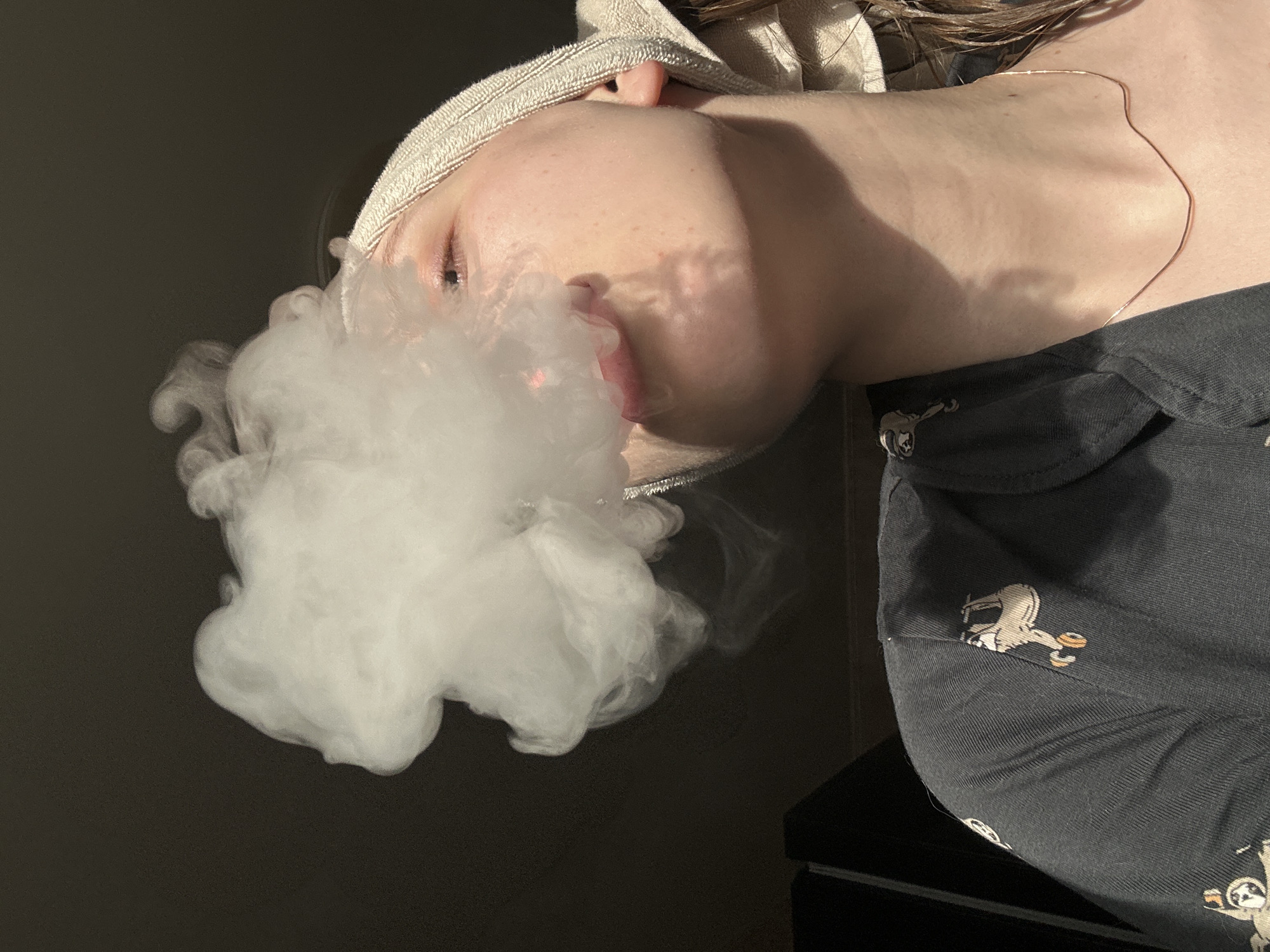 The image presents a close-up, angled view of a person exhaling a thick cloud of white vapor from their mouth. Only the lower half of the subject's face is clearly visible, including the mouth, chin, and a portion of one eye. A beige, textured fabric, possibly a towel or blanket, is draped over the head.
A slender, light-colored necklace rests on the person's chest and neck. The individual is wearing a dark-colored garment, likely a t-shirt, adorned with light-colored, stylized patterns, one depicting a figure with a helmet.
Strong, directional light illuminates the face and vapor, creating distinct shadows and suggesting either direct sunlight or a focused artificial light source. The background is dark and indistinct, indicating an indoor setting or a deeply shaded area. The image is rotated clockwise, placing the person's head towards the bottom left. The expression appears neutral. There is no visible text. This scene is set in Chaikovsky, Russia.