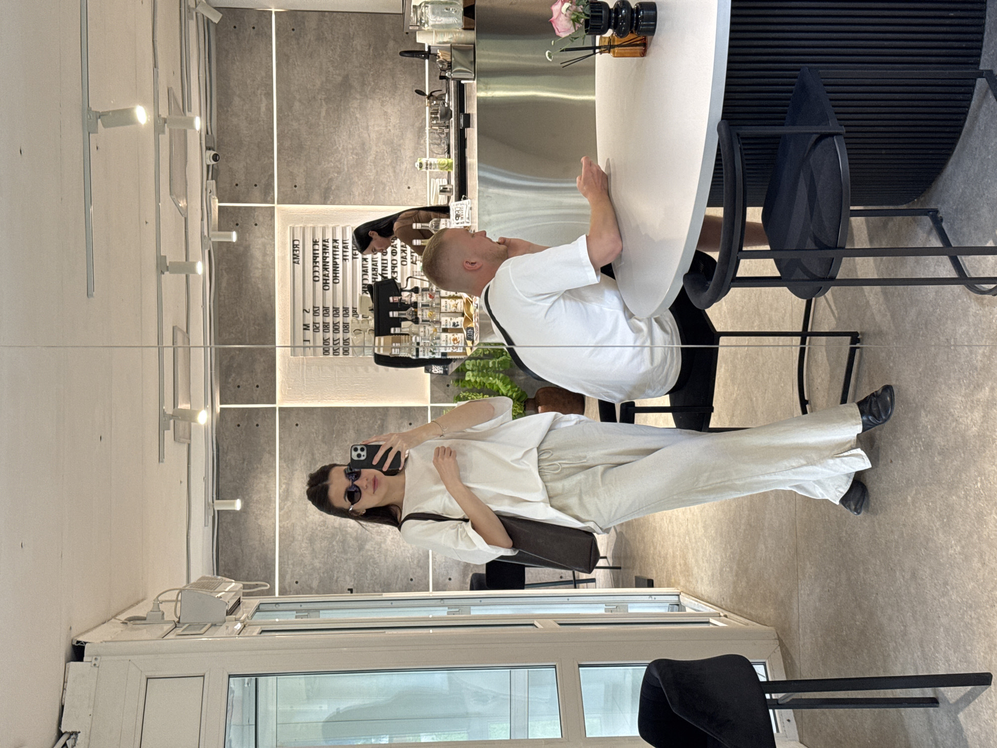 In an indoor cafe in Chaikovsky, Russia, a young woman with dark hair and sunglasses takes a selfie in a mirror. She is wearing a white t-shirt and light-colored linen pants, carrying a dark tote bag over her left shoulder. To her right, a man with blond hair wearing a white t-shirt sits at a round table, looking at his phone. Behind them, a barista with dark hair in a brown top works behind a counter stocked with bottles and equipment. The cafe features concrete walls, track lighting, and modern furniture, including black bar stools and a round table with a fluted base. A potted plant and a small floral arrangement are visible on the table.