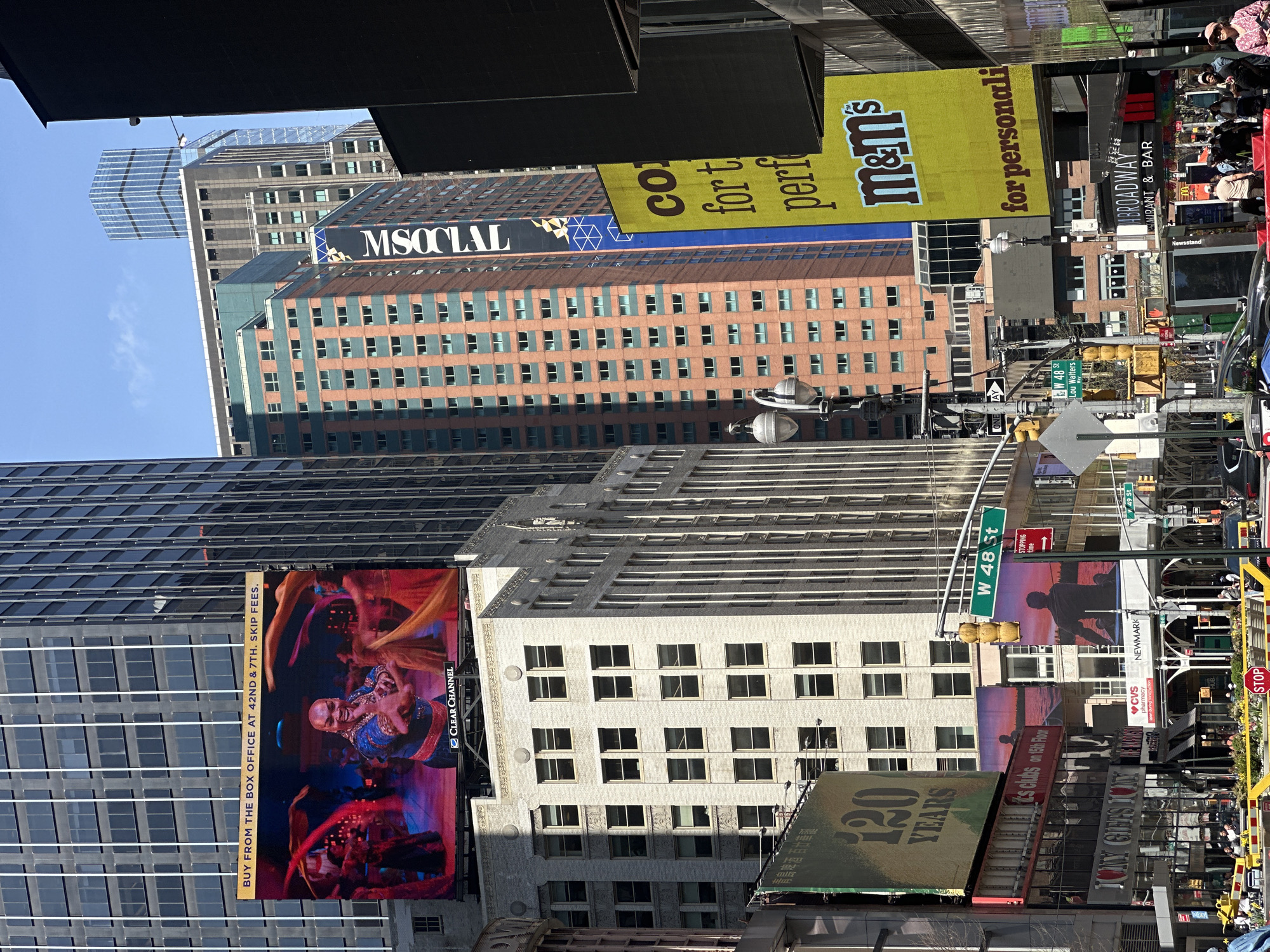 This photograph captures a bustling street scene in the heart of Times Square, New York City. Dominating the view are towering buildings adorned with numerous advertisements and billboards. Among the recognizable signs are promotions for "M&M's," "MSOCIAL," and a theatrical show advertised on a large digital billboard, encouraging viewers to "BUY FROM THE BOX OFFICE AT 42ND & 7TH. SKIP FEES." Street signs indicate the location as West 48th Street, further pinpointing the scene's location. Traffic lights and pedestrian signals are visible, along with dense pedestrian traffic, indicating a lively urban environment. There is a banner celebrating "120 YEARS." Various other smaller signs advertise restaurants and businesses.