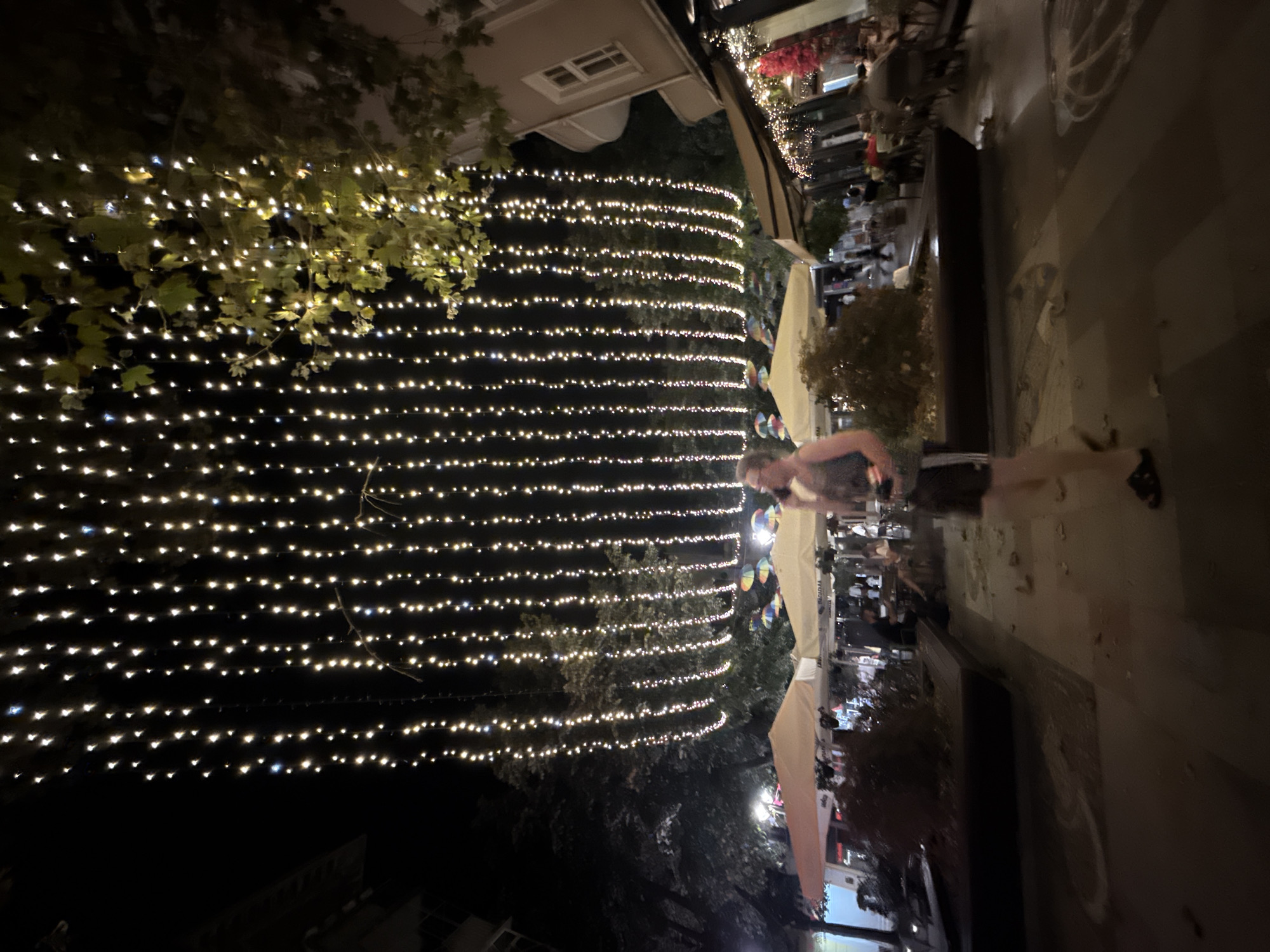 The image captures a night scene on a street in Burgas, Bulgaria, adorned with numerous strings of small, bright lights hanging overhead. In the foreground, a person is walking and talking on a mobile phone. The background reveals an outdoor seating area with umbrellas and tables, likely a cafe or restaurant, with other patrons present. Trees with green leaves are visible, illuminated by the string lights. The overall ambiance suggests a lively evening in an urban setting.