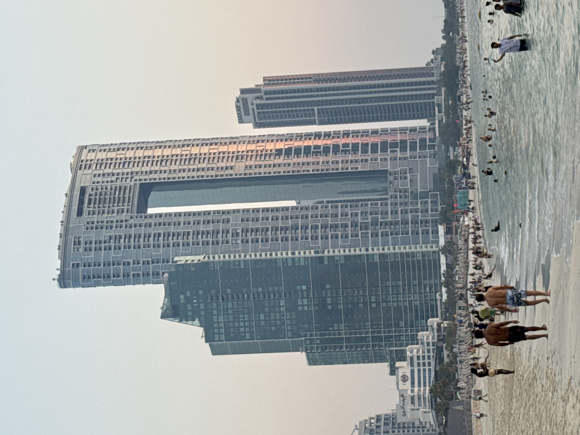 The image shows a crowded beach scene with a backdrop of modern skyscrapers in Dubai, United Arab Emirates. In the foreground, several people are walking on the sand and wading in the shallow water. The sea is calm and appears to be the Persian Gulf.
Dominating the background are several tall buildings, characteristic of Dubai's skyline. The most prominent is a very tall skyscraper with a distinctive rectangular opening in the upper section, reflecting the warm hues of what appears to be a sunset or sunrise. To its left, another large, dark glass skyscraper is visible. To the right of the main skyscraper, a slightly shorter, slender building with the "EMAAR" logo at its top stands out. Further in the distance, more buildings and structures line the coastline, indicating a densely developed urban area.
The beach itself is populated with many people, some relaxing on lounge chairs under umbrellas, while others are swimming or walking along the shore. The overall atmosphere suggests a popular recreational area within the city, likely a tourist destination or a residential beachfront. The sky is a pale, hazy blue, common in coastal areas.