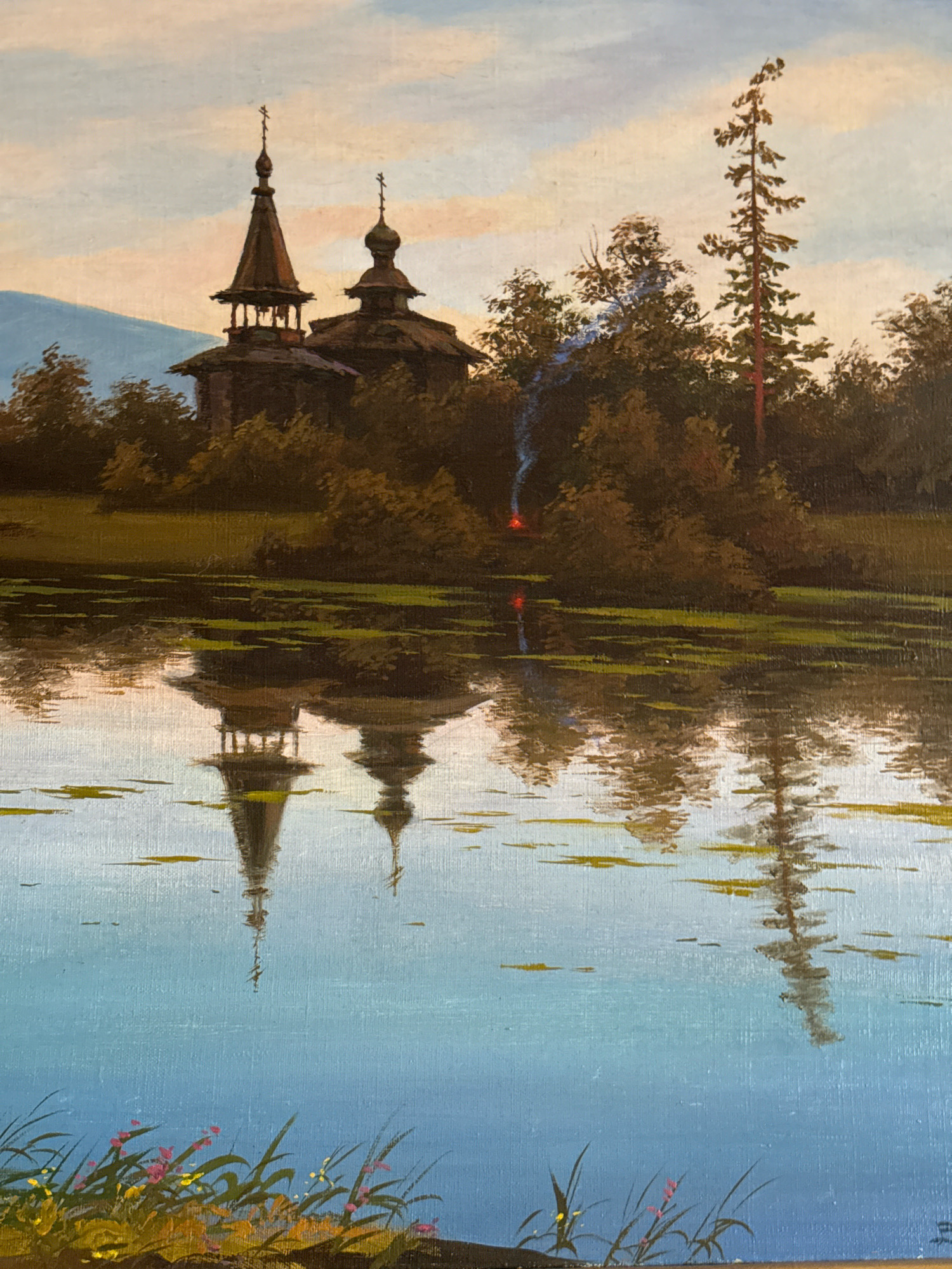 This is a painting depicting a serene landscape scene. In the background, a wooden church with multiple onion domes and crosses stands amidst lush green trees. A thin plume of blue and orange smoke rises from behind the church. To the right of the church, a tall, slender evergreen tree stands out against a pastel-colored sky. In the foreground, a calm body of water reflects the church, trees, and sky, creating a mirrored image. Lily pads are scattered across the surface of the water. Along the bottom edge of the painting, a patch of wildflowers and grasses adds a touch of vibrant color to the scene. The painting captures a peaceful, possibly rural, setting with architectural elements and natural beauty. There is no indication of the city of Krasnoyarsk, Russia, in the image.