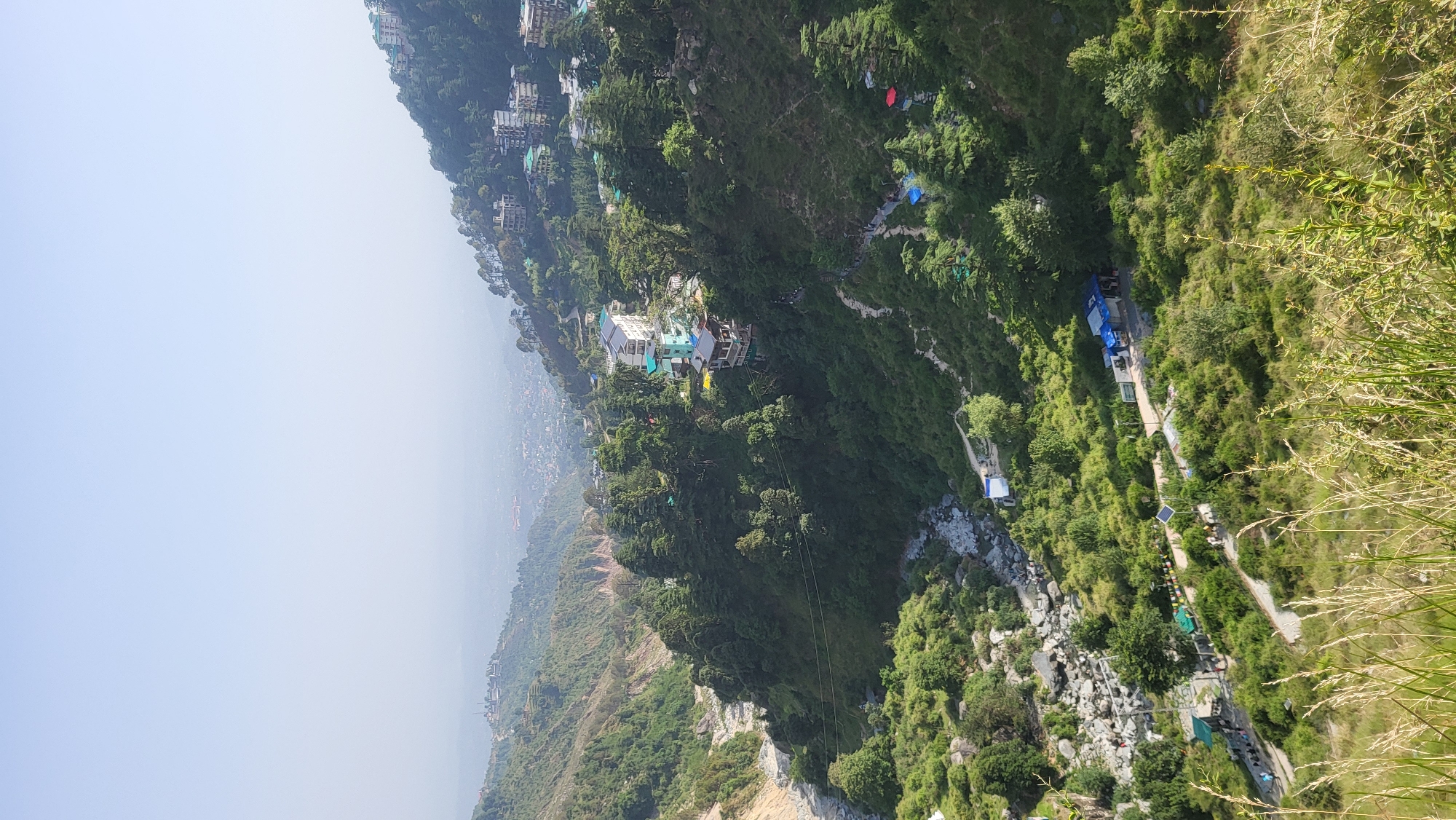 The image depicts an aerial view of Bhagsu, India, showcasing a steep hillside densely covered with lush green vegetation. Buildings are scattered along the upper slopes, indicating residential or commercial development. A prominent white structure with blue accents is situated midway down the slope. A river or stream cuts through the landscape, marked by visible water flow and rocky terrain. A trail or path runs alongside the water, punctuated by tents or structures, suggesting a recreational area or campsite. The sky is overcast, casting a hazy light over the scene. Tall grasses are visible in the foreground, indicating the viewpoint location.