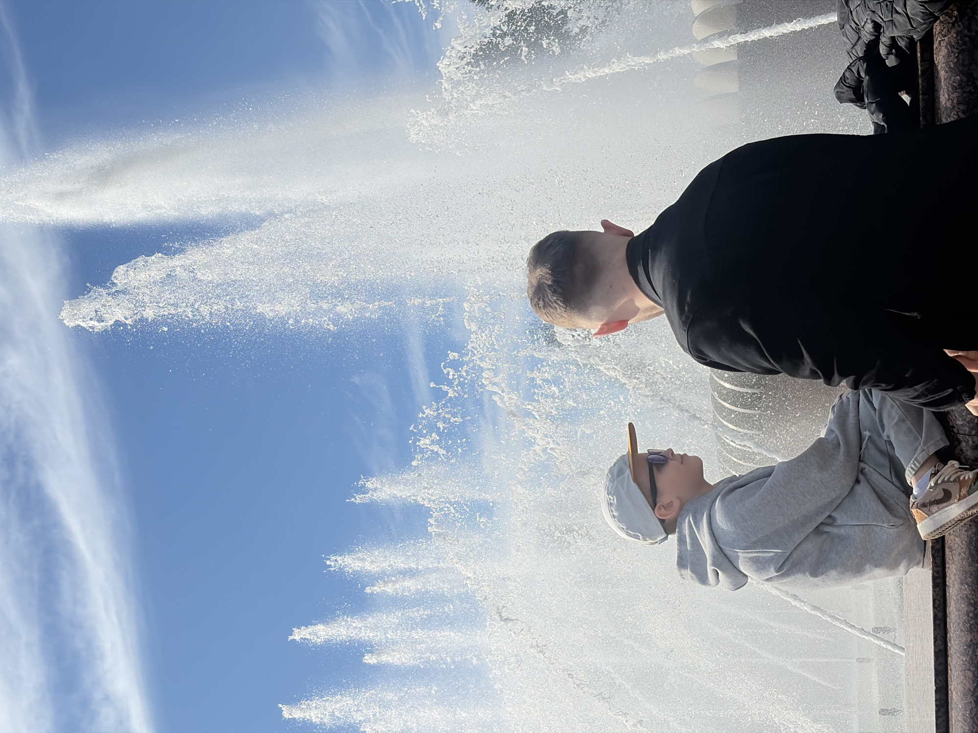 The image depicts two individuals, an adult male and a child, observing a powerful fountain under a clear blue sky with scattered wispy clouds. The original image is oriented vertically, showing the subjects looking up at the fountain's spray.
The adult male, dressed in a black long-sleeved shirt, is positioned behind and slightly above the child, with his back partially to the viewer, looking upwards towards the fountain. The child is wearing a light grey hooded sweatshirt, a white baseball cap, and sunglasses. Their right foot is visible, wearing a sneaker with light brown and white accents. The child appears to be seated or leaning on a dark, possibly granite or concrete, edge of the fountain structure, also looking towards the water.
Multiple strong jets of water from the fountain are spraying high into the air, creating a dramatic and misty spectacle against the bright sky. The sunlight illuminates the water, causing it to sparkle. The background consists primarily of the expansive blue sky and the white froth of the fountain's spray.
The scene takes place during bright daylight, suggesting good weather conditions. The setting appears to be a public area or park where a large fountain is a prominent feature, consistent with locations found in Saint Petersburg, Russia. There is no visible text in the image.
