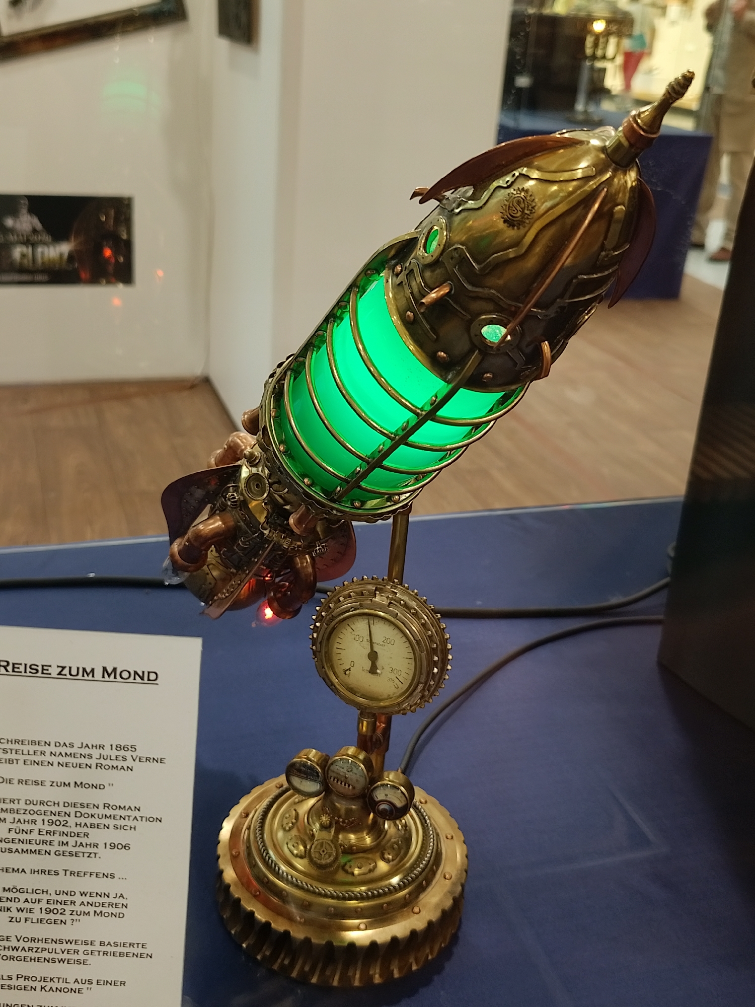 A steampunk-style lamp in the shape of a rocket sits on a blue surface. The lamp is made of brass-colored metal and has a green illuminated cylindrical section. The base is a circular structure with gears and gauges. To the left of the lamp is a white paper with German text, titled "REISE ZUM MOND" (Journey to the Moon).