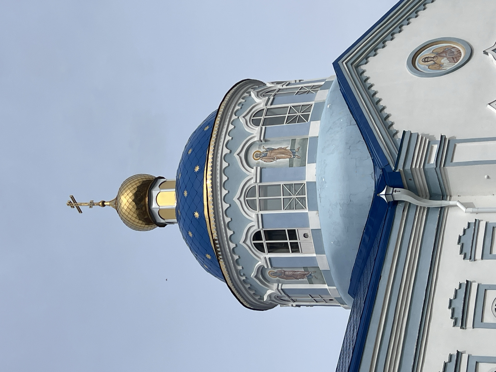 The image captures a section of the Church of the Nativity of Christ, located in Kargopolye, Russia. The view is from below, looking upward at the church's architecture. A blue dome, adorned with golden stars and topped with a golden onion dome and a cross, is prominent. Below the blue dome is a light blue structure with arched windows and religious paintings of figures. To the right, a section of the church's facade is visible, featuring white walls with light blue accents and a circular painting within. The sky is a pale blue.